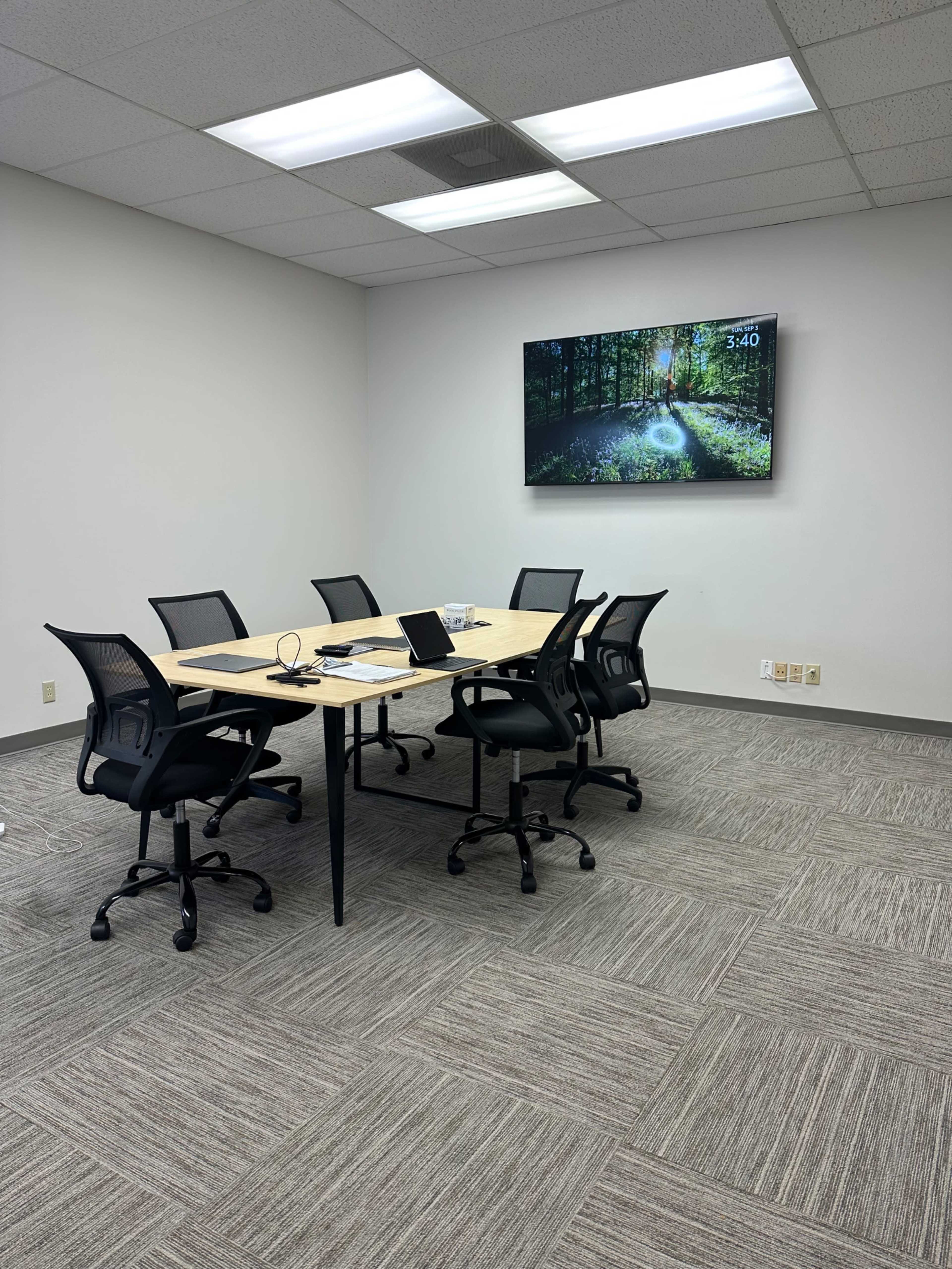 A modern conference room features a rectangular table with six black chairs, a laptop, and a television displaying a forest scene on the wall.