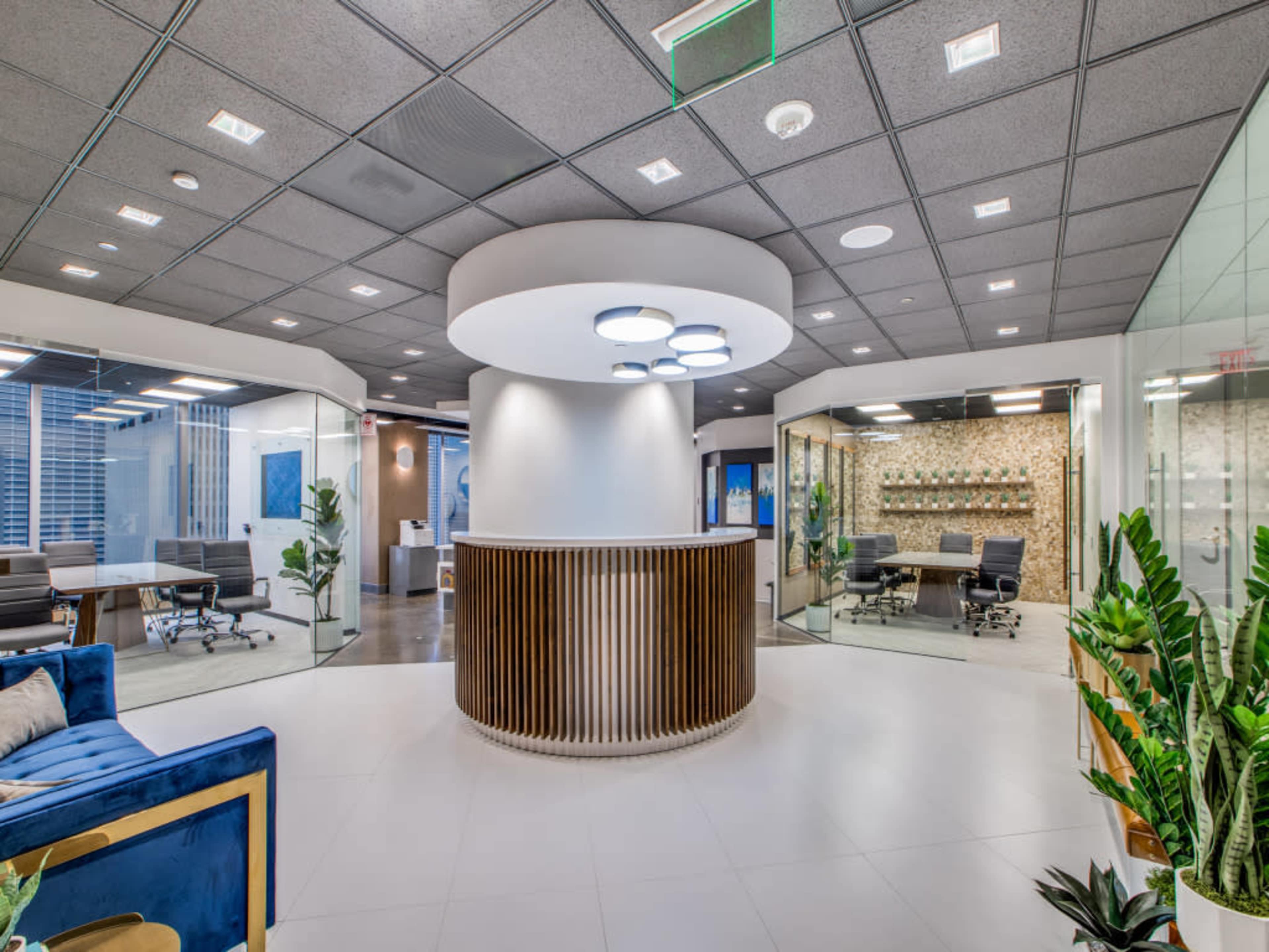 A modern office lobby with a circular reception desk, glass meeting rooms, and a seating area featuring a blue couch and indoor plants.