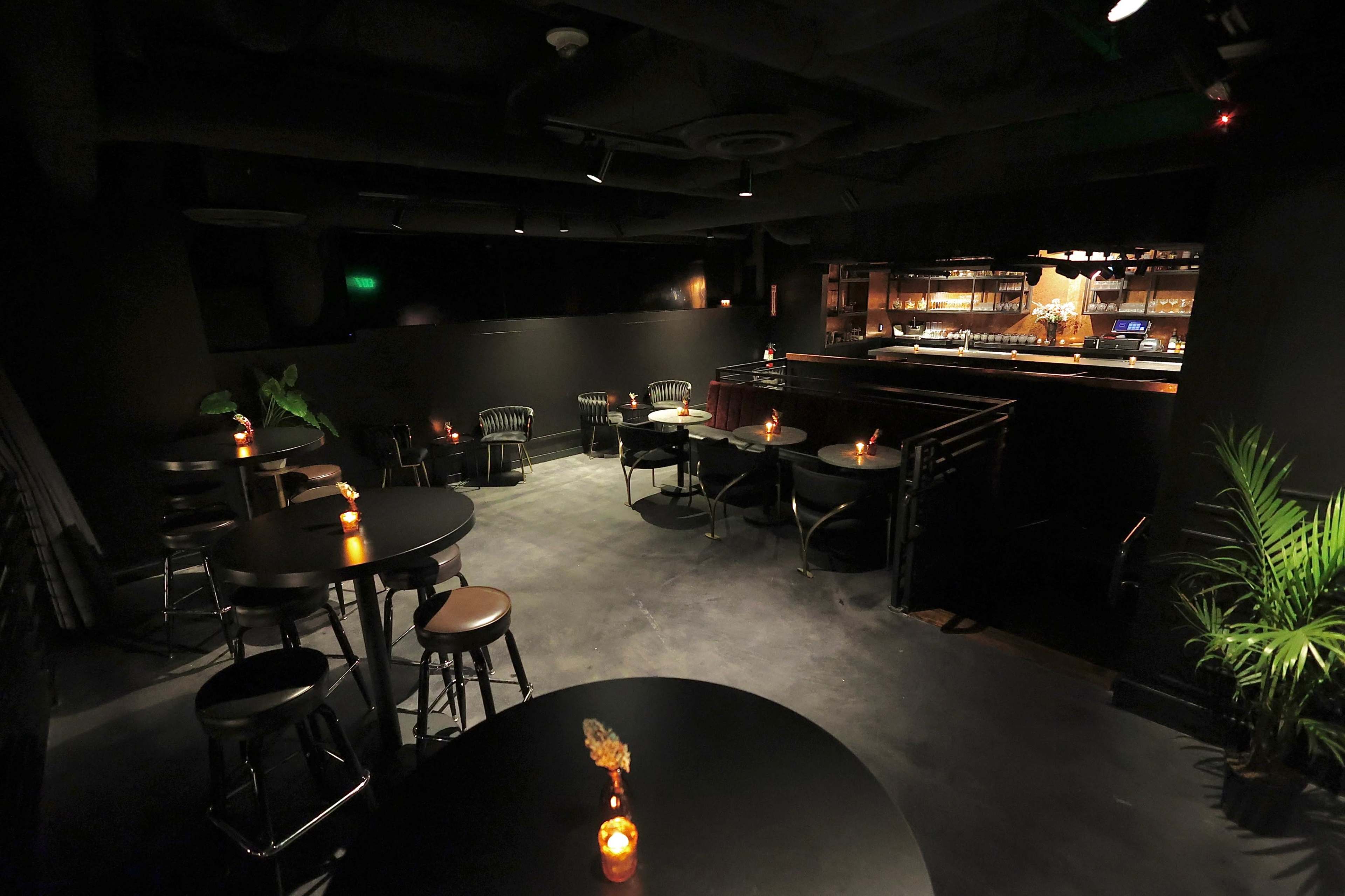 The image shows a dimly lit bar with black walls, tables, and chairs, featuring small candles on each table and a bar at the back.