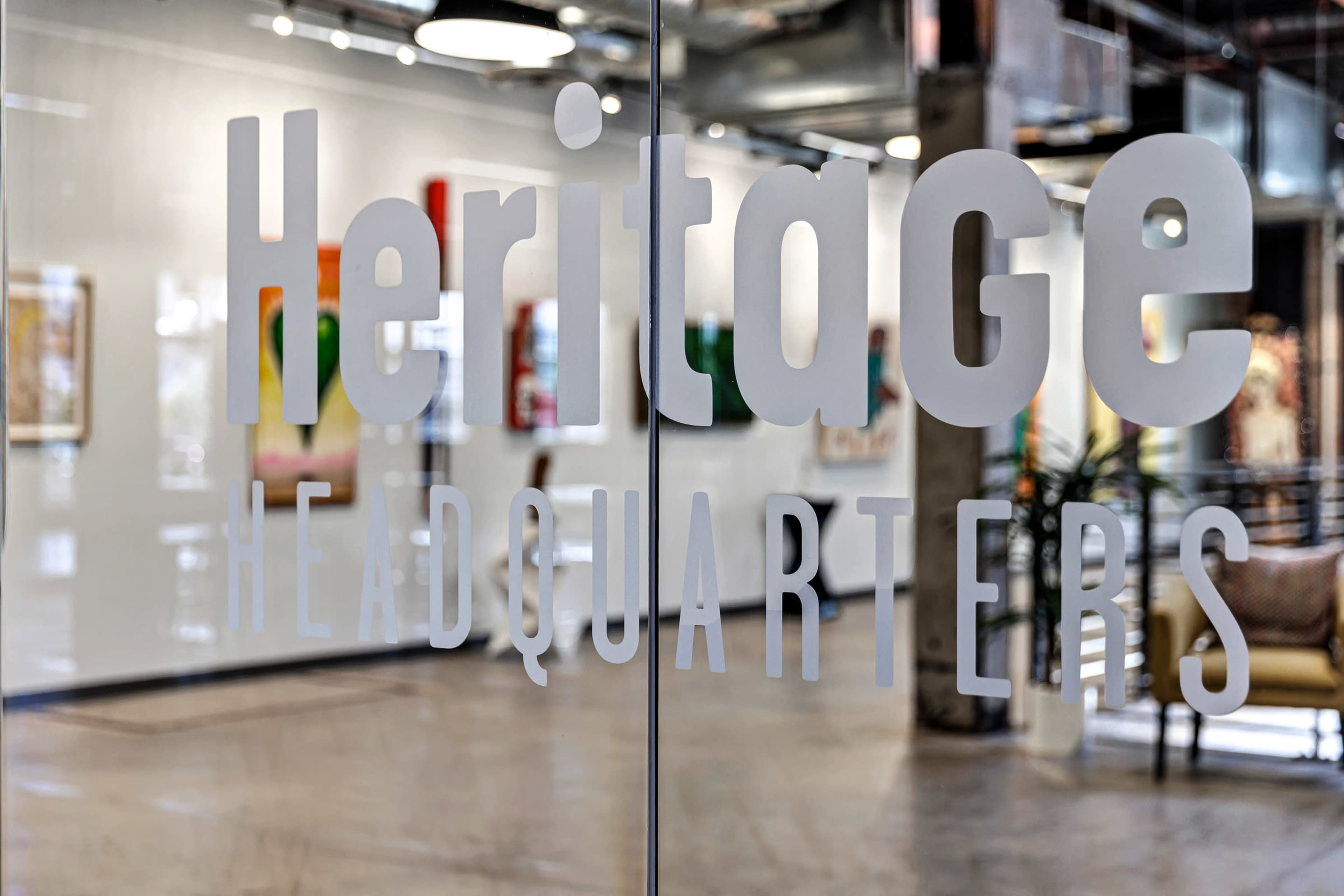 The image shows the entrance of a building with the words "Heritage Headquarters" prominently displayed on a glass door, revealing a bright art-filled interior.