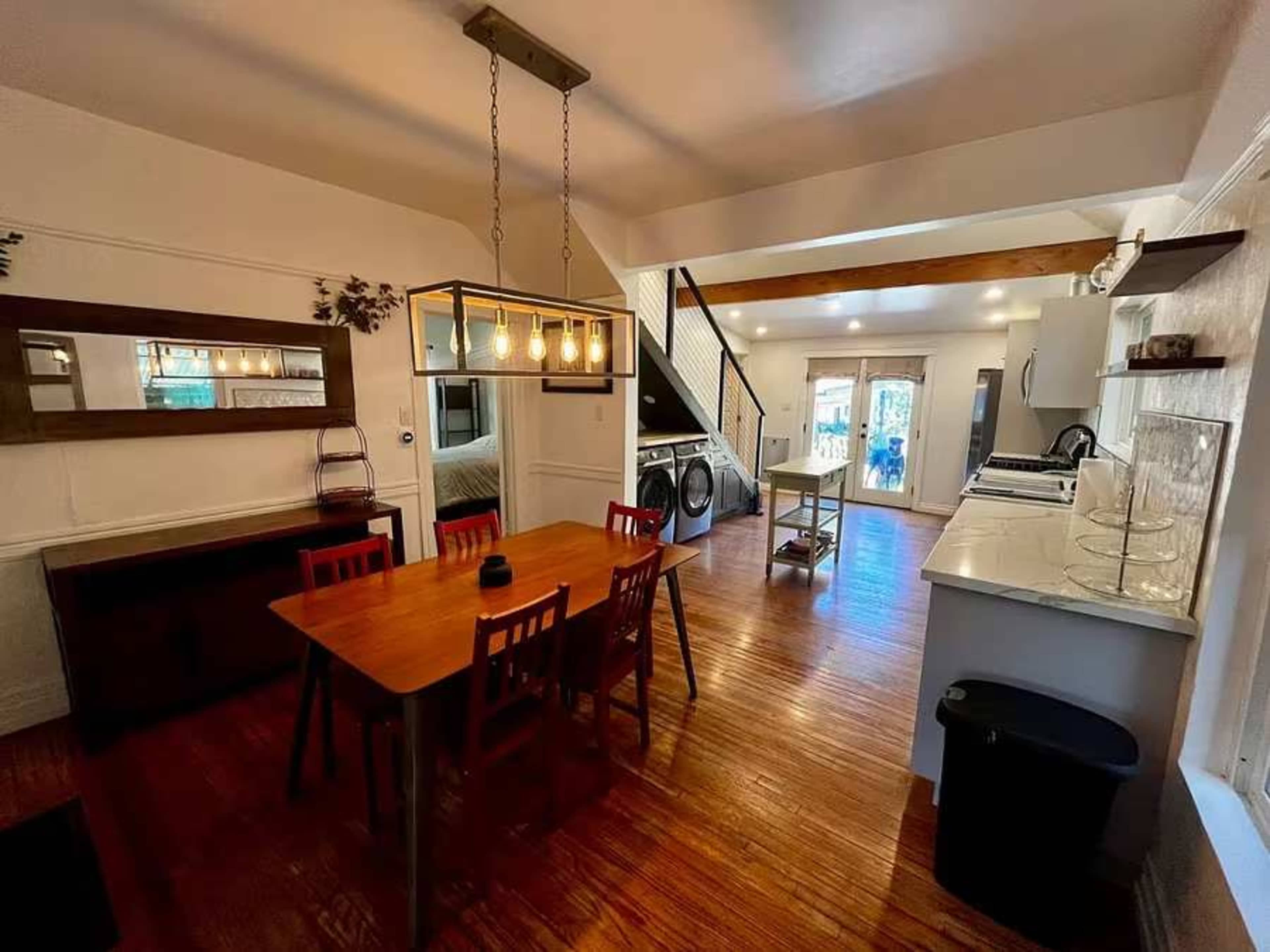 The image shows a spacious dining area with a wooden table and chairs, connected to an open kitchen illuminated by pendant lighting.