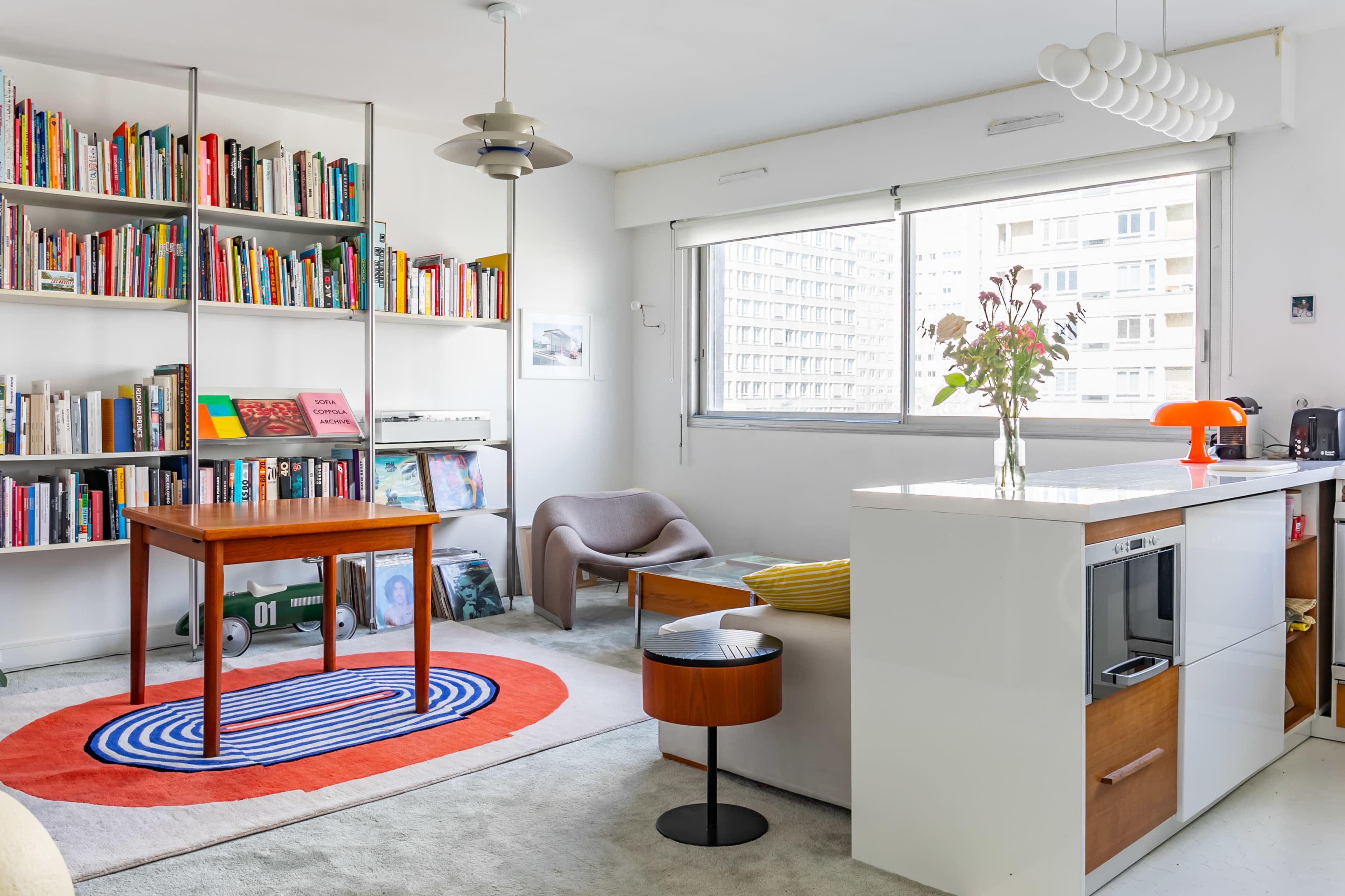 A modern living space features a bookshelf filled with books, a wooden table, a chair, a decorative rug, and a kitchen area with cabinetry.