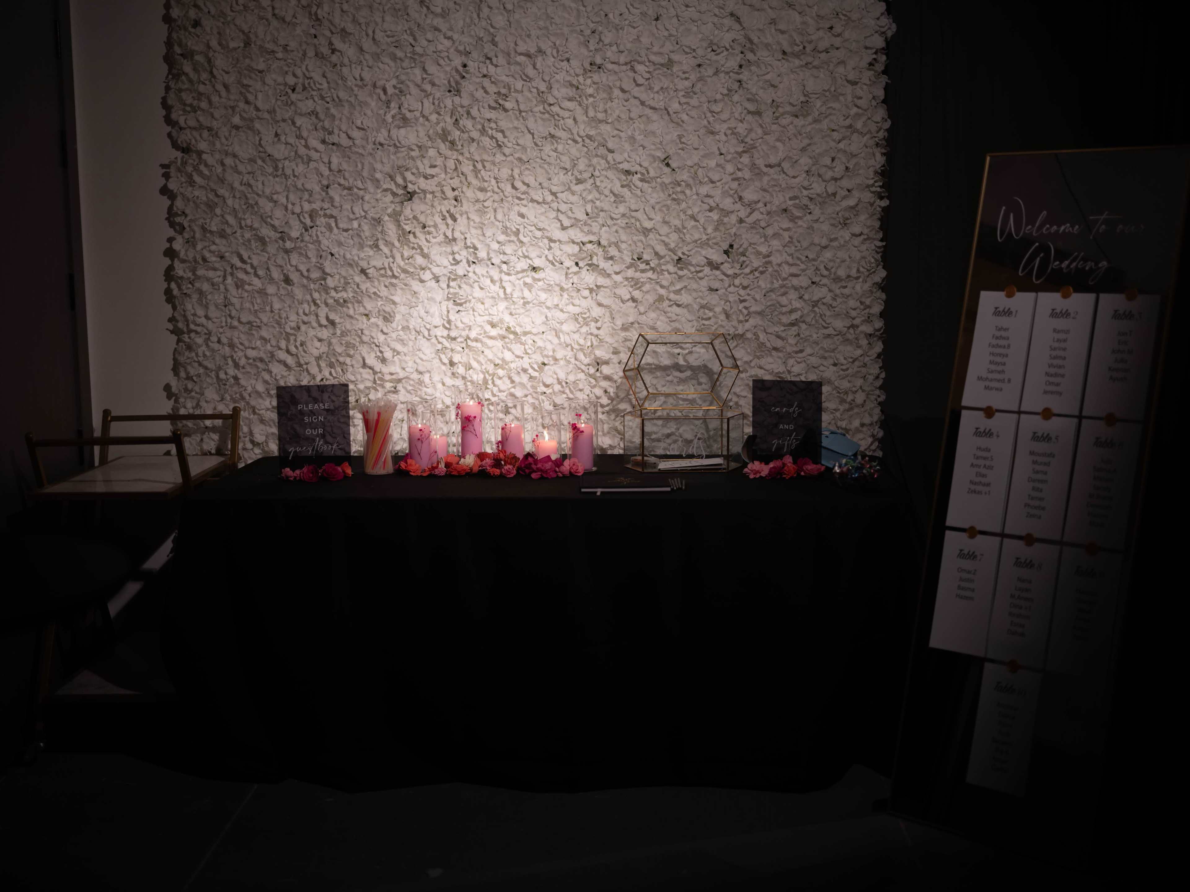 A table is set against a textured wall, adorned with candles, decorative flowers, and a sign welcoming guests, alongside a list of names.