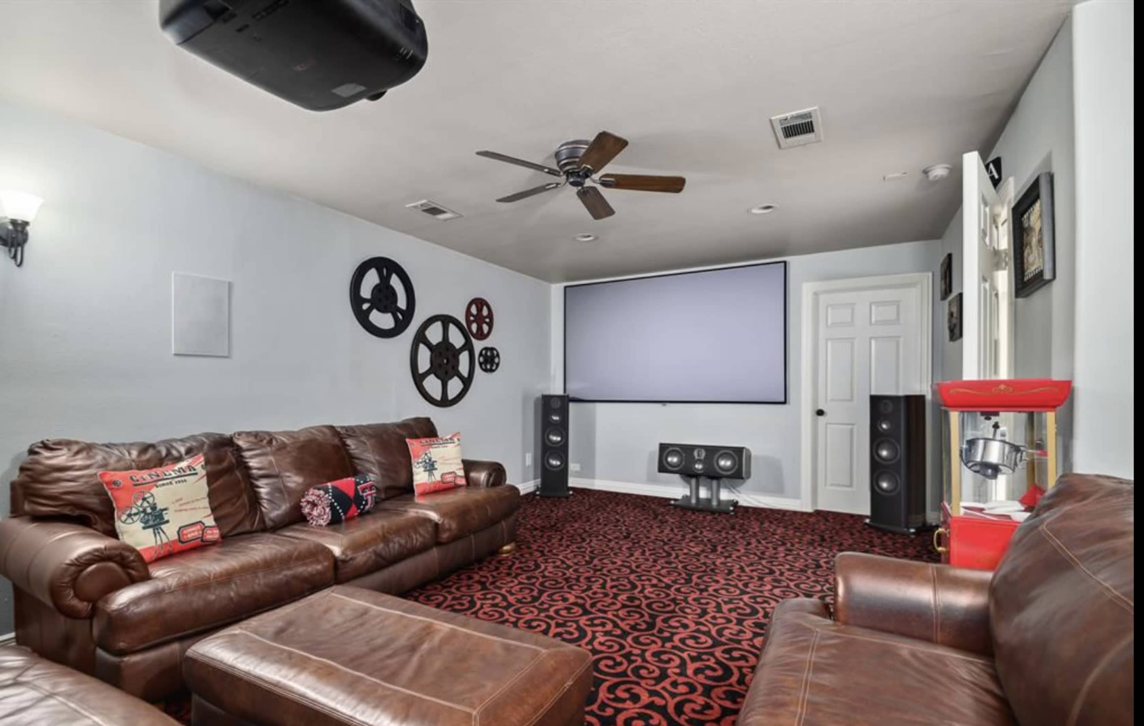 A home theater room featuring brown leather couches, a large projection screen, and speaker systems against a patterned carpet.