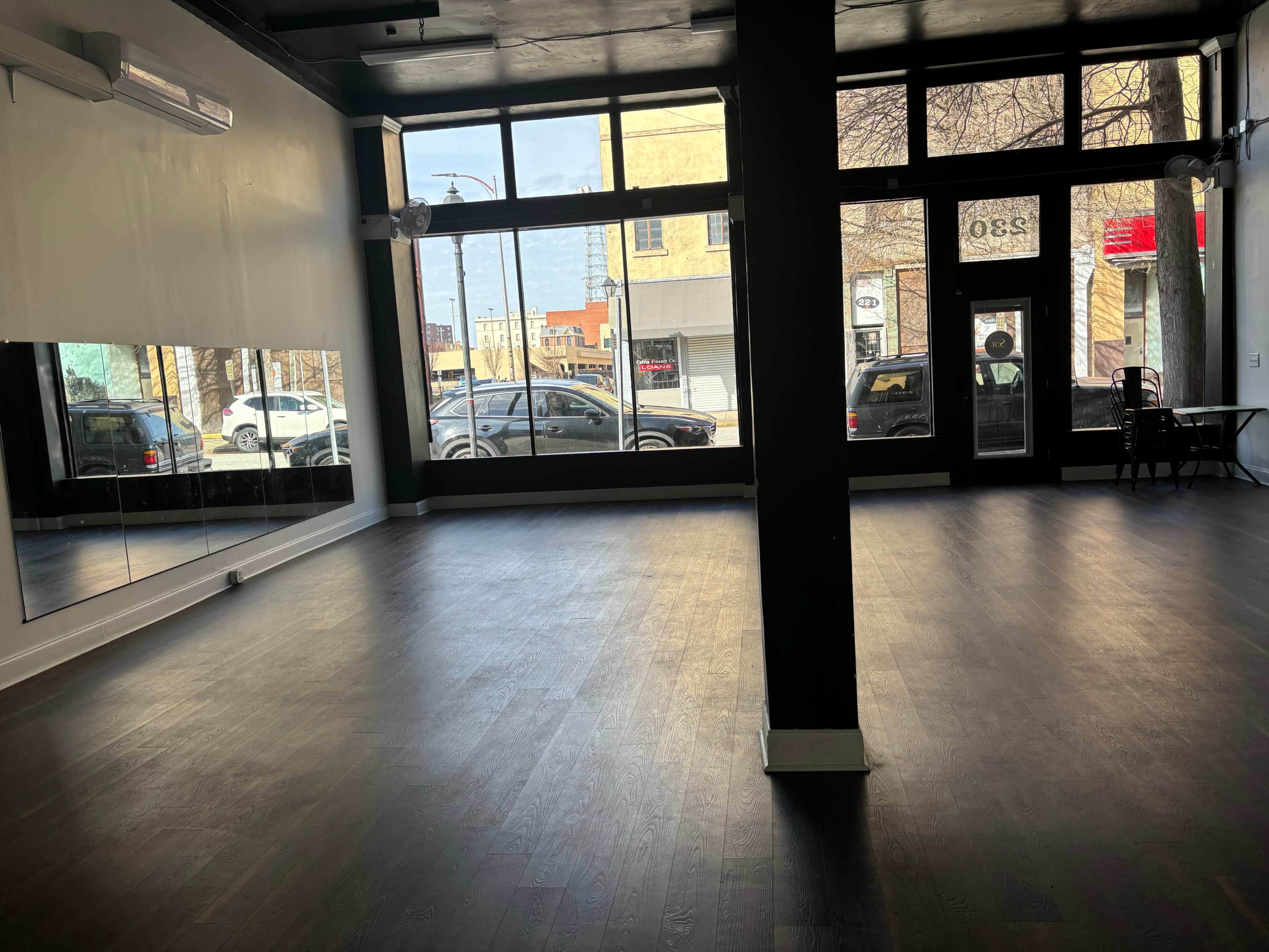 An empty interior space with hardwood floors, large windows letting in natural light, and a wall mirror.