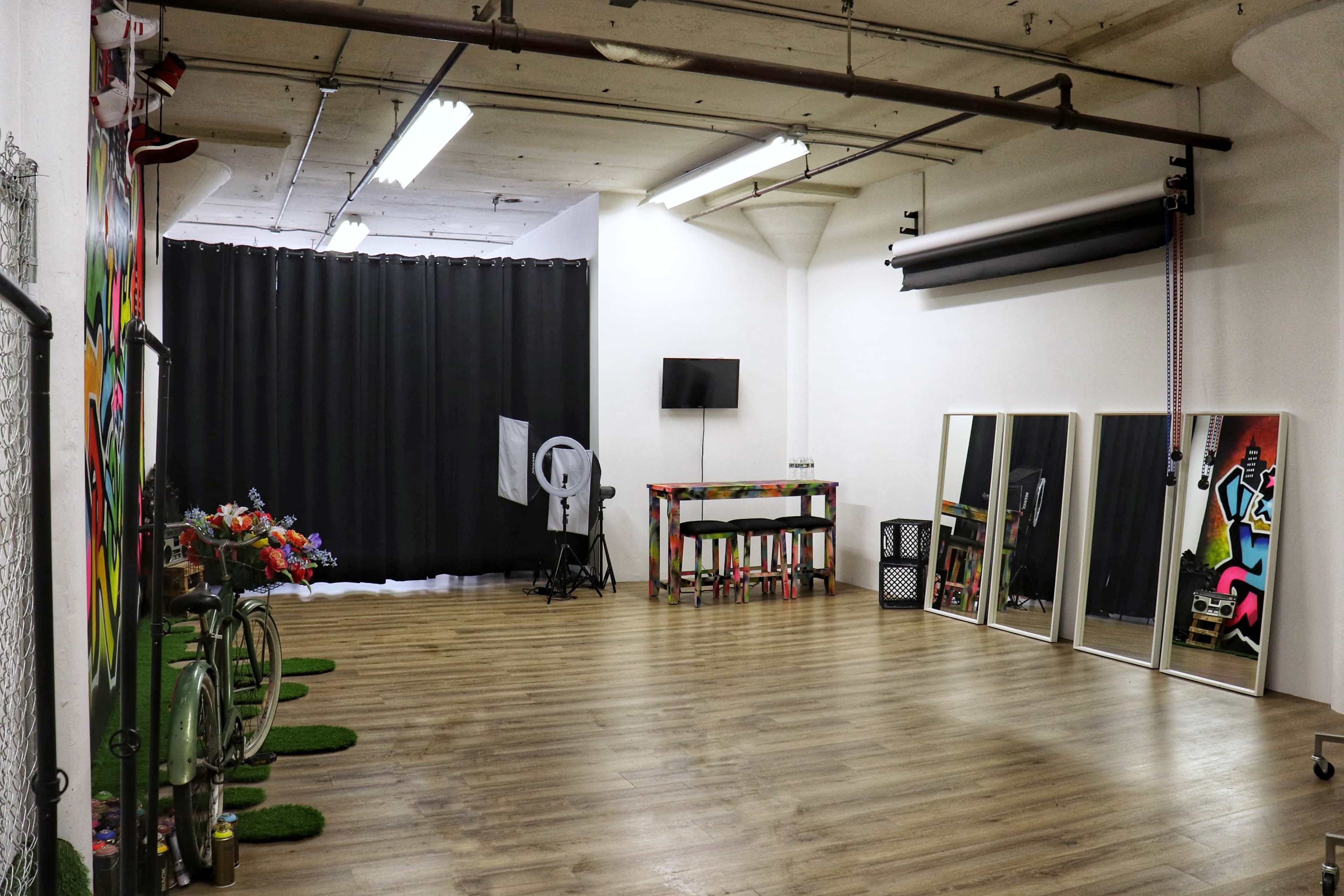 A spacious, brightly lit room with wooden flooring, featuring a black curtain on one side, a wall-mounted TV, a table with colorful decorations, and two large mirrors.