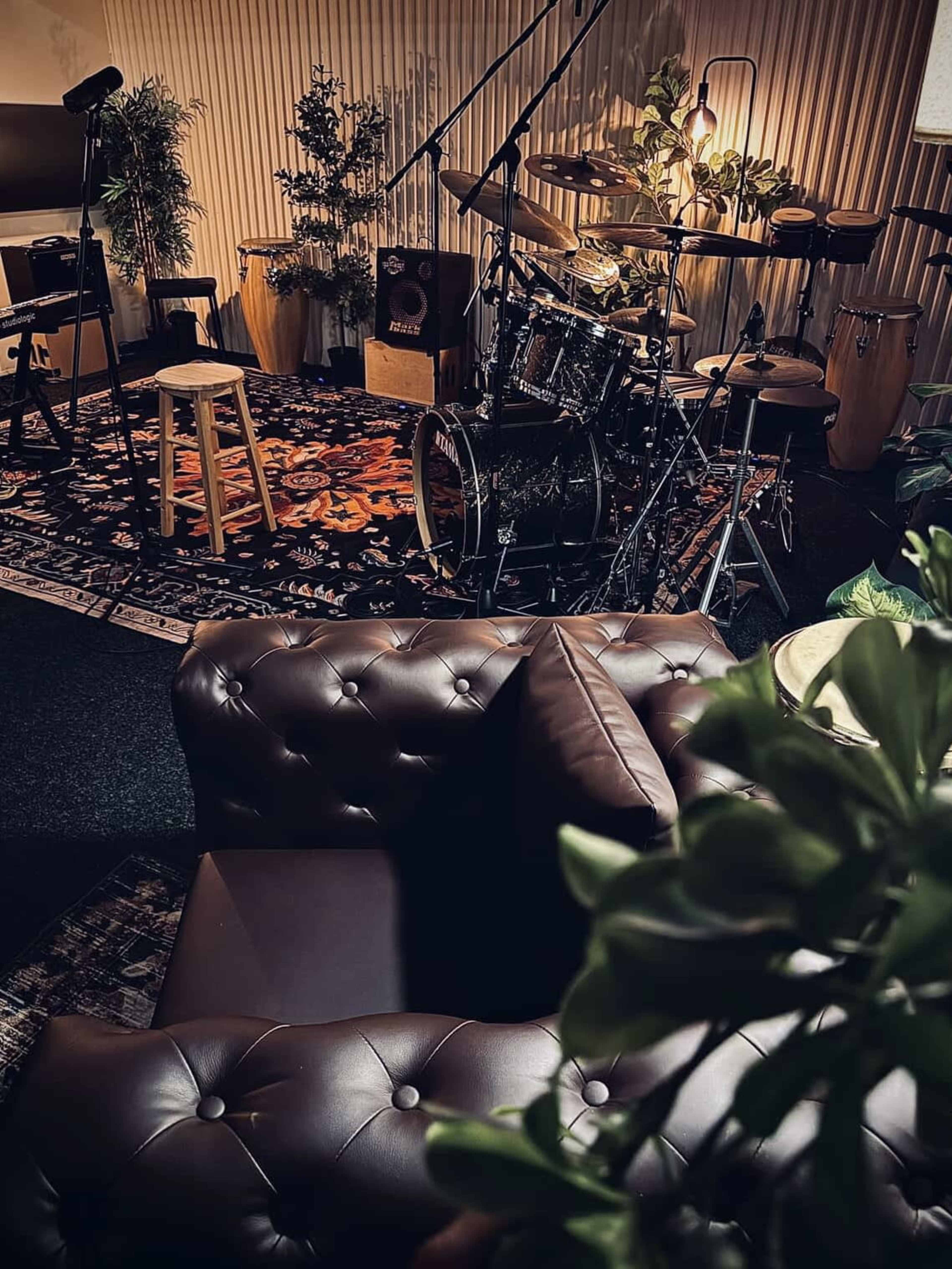 The image shows a cozy music studio setup with a drum kit, various instruments, a stool, and a dark leather couch surrounded by plants on a patterned rug.