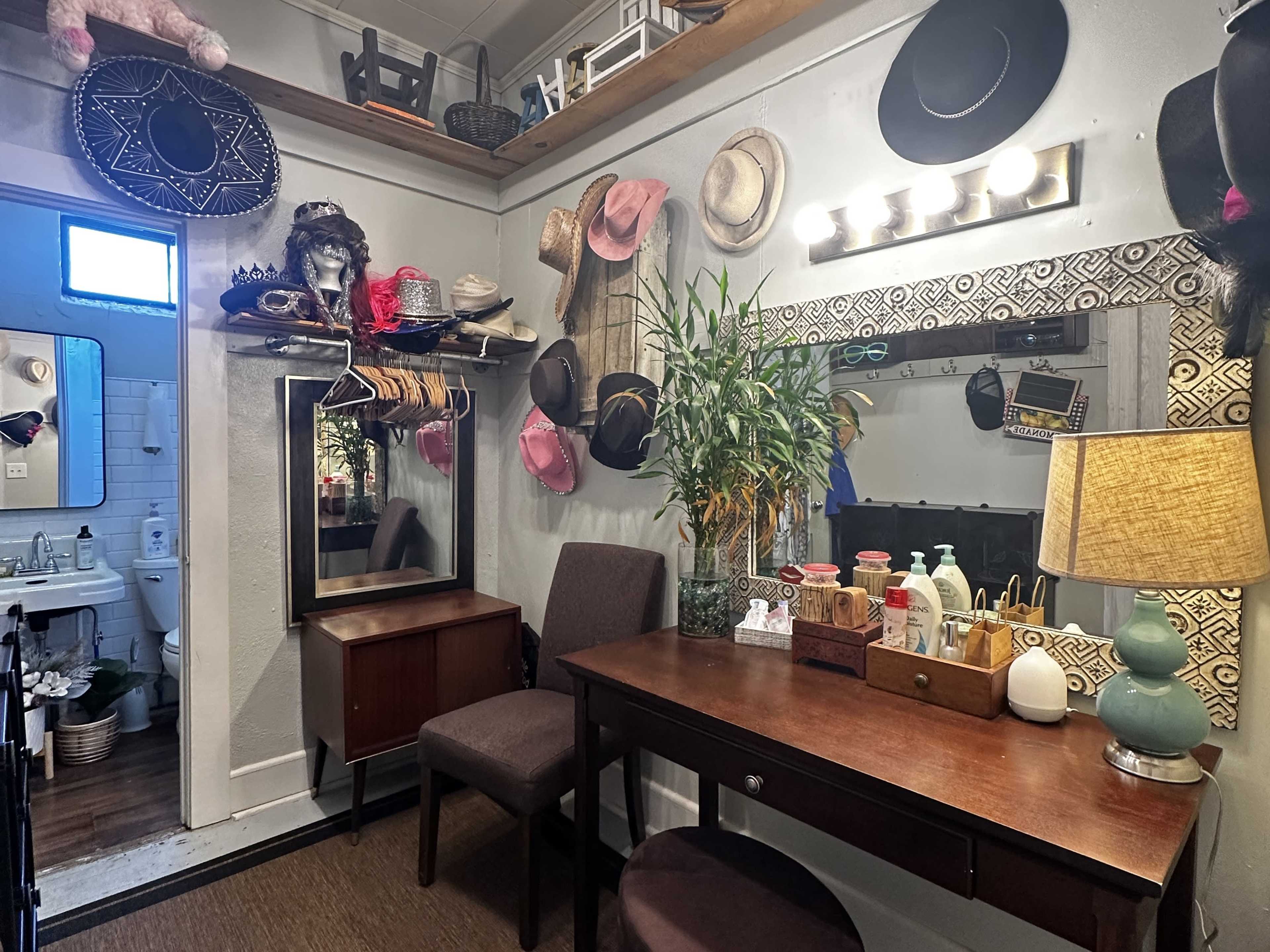 The scene features a small dressing area with a mirror, two chairs, a table, a clothing rack, and various hats hanging on the walls.