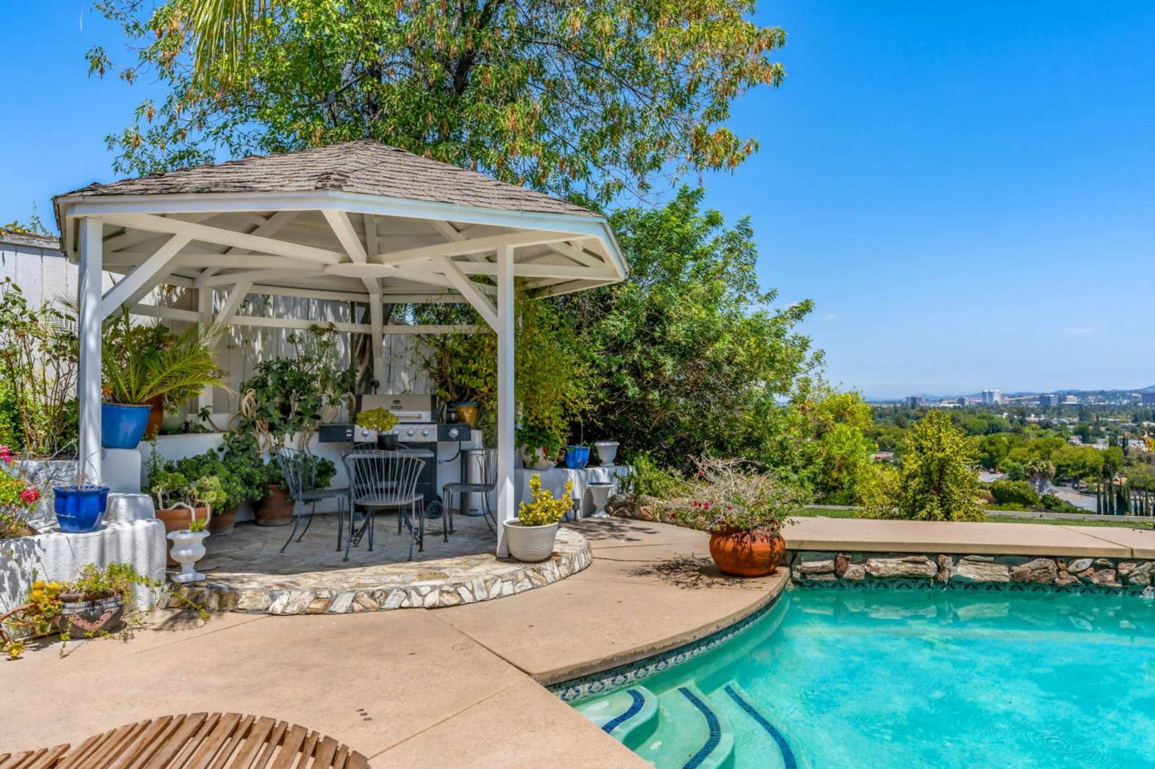 A gazebo with seating overlooks a swimming pool, surrounded by landscaped greenery and distant city views.