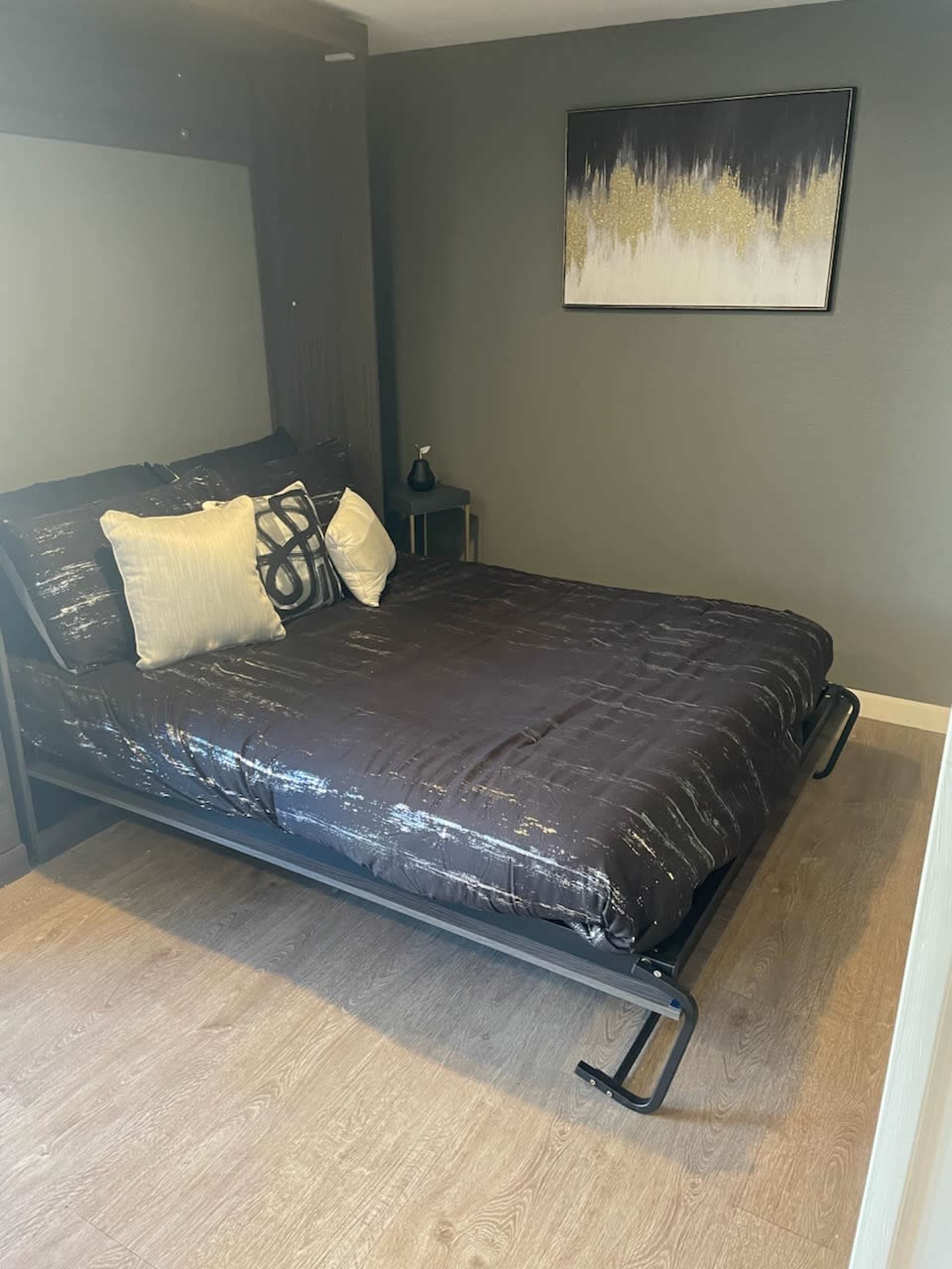 A bed with a black and silver comforter is positioned against a neutral wall in a neatly arranged bedroom.