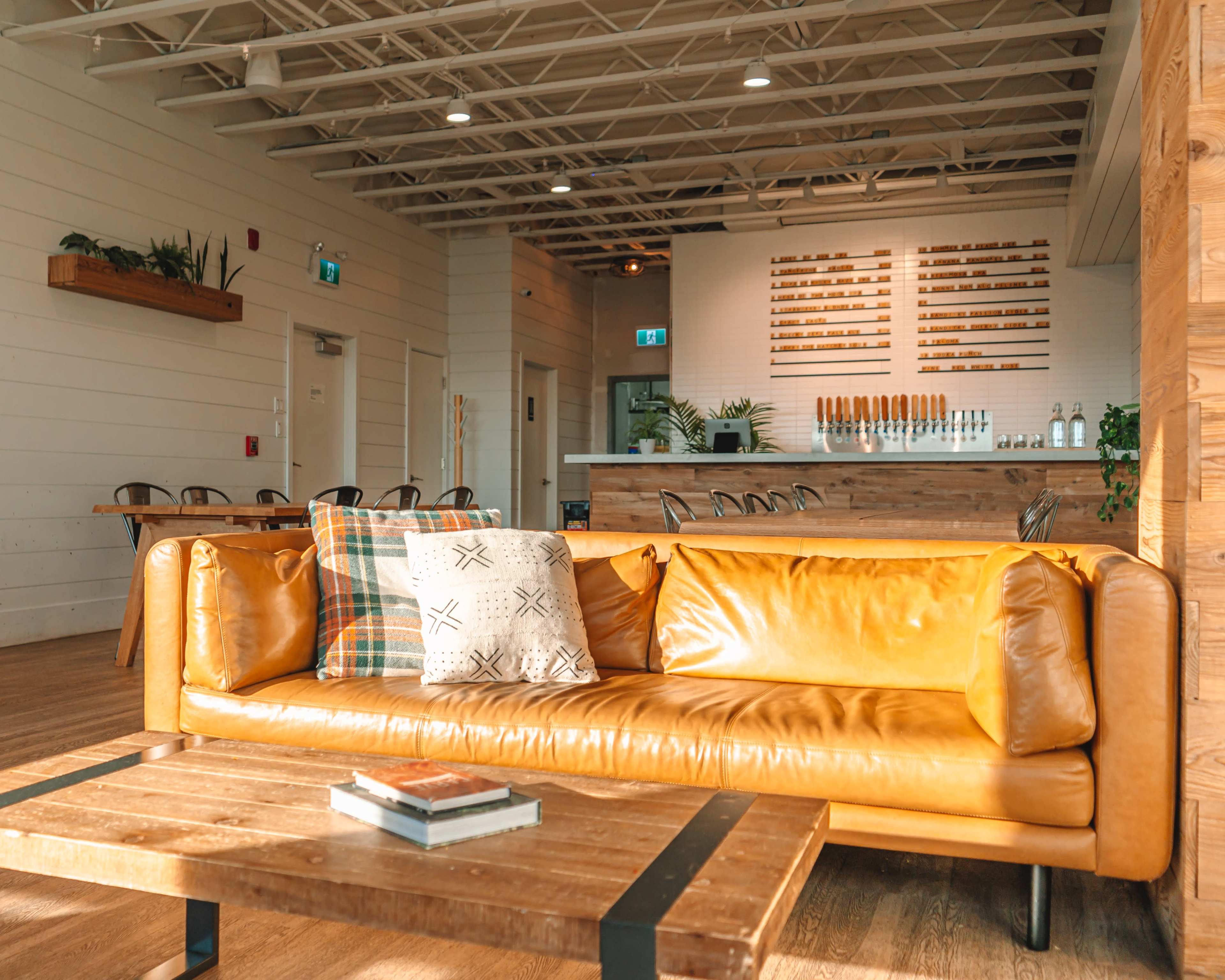 A cozy interior of a café featuring a brown leather sofa, a wooden coffee table, and a bar area with multiple beer taps in the background.