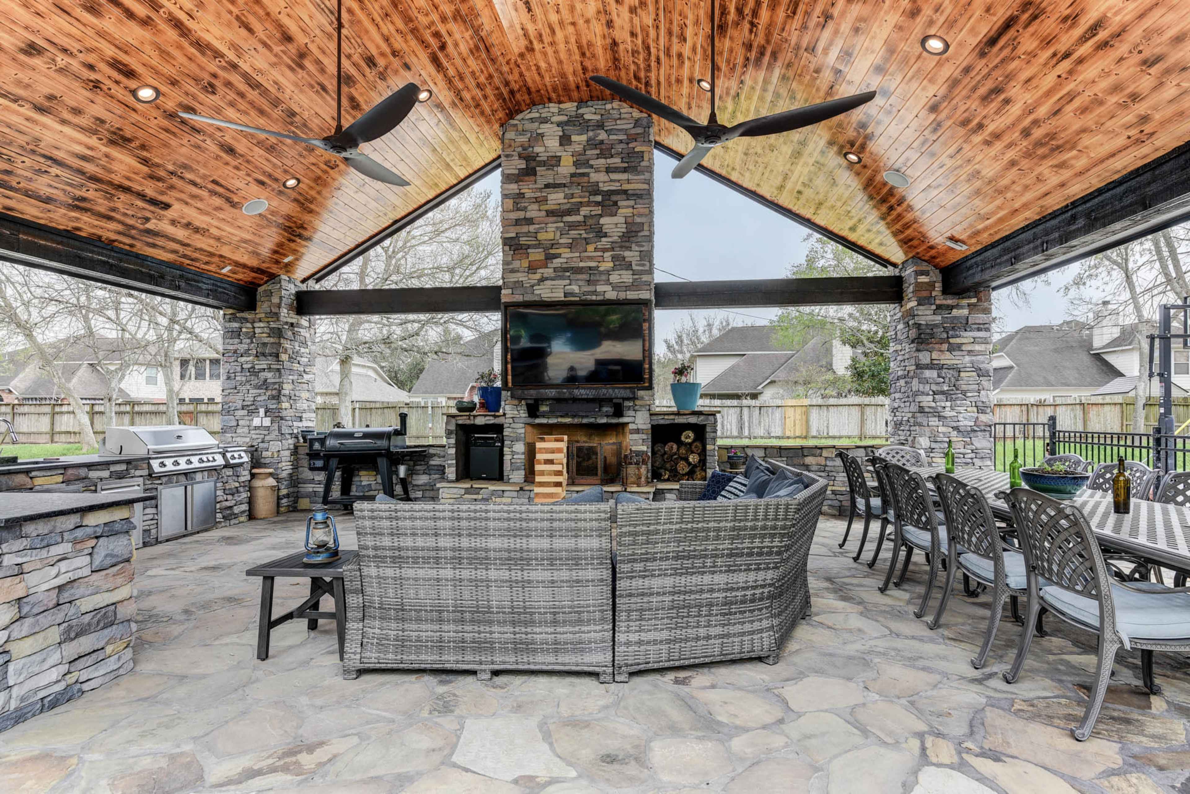 A spacious outdoor patio featuring a stone fireplace, modern cooking appliances, and a seating area with wicker furniture under a wood-paneled ceiling.