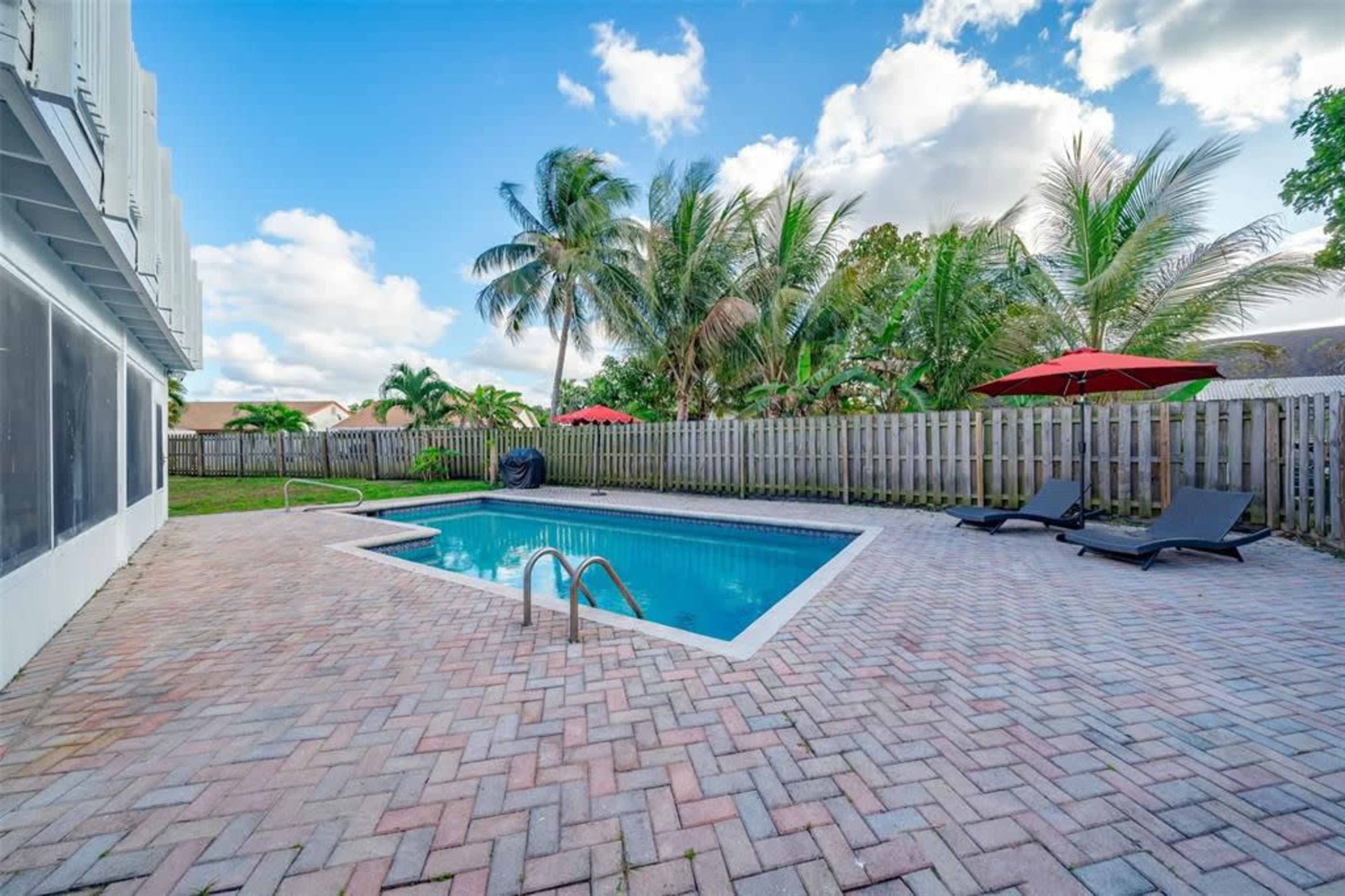 A rectangular swimming pool is surrounded by a paved patio with two lounge chairs and palm trees, under a partly cloudy blue sky.