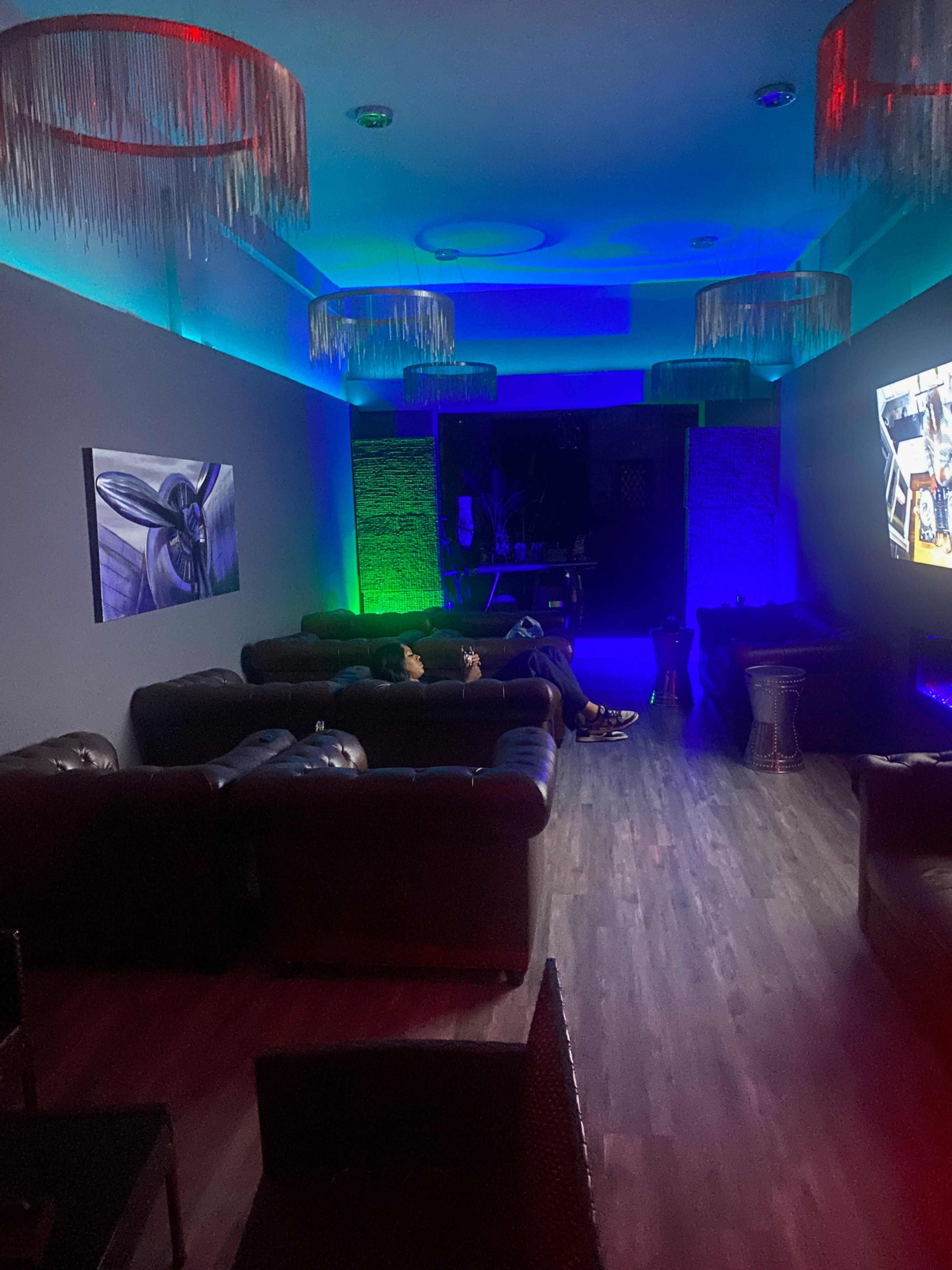 Two people sit on dark leather sofas in a dimly lit room with colorful ambient lighting, facing a large screen that displays a video.