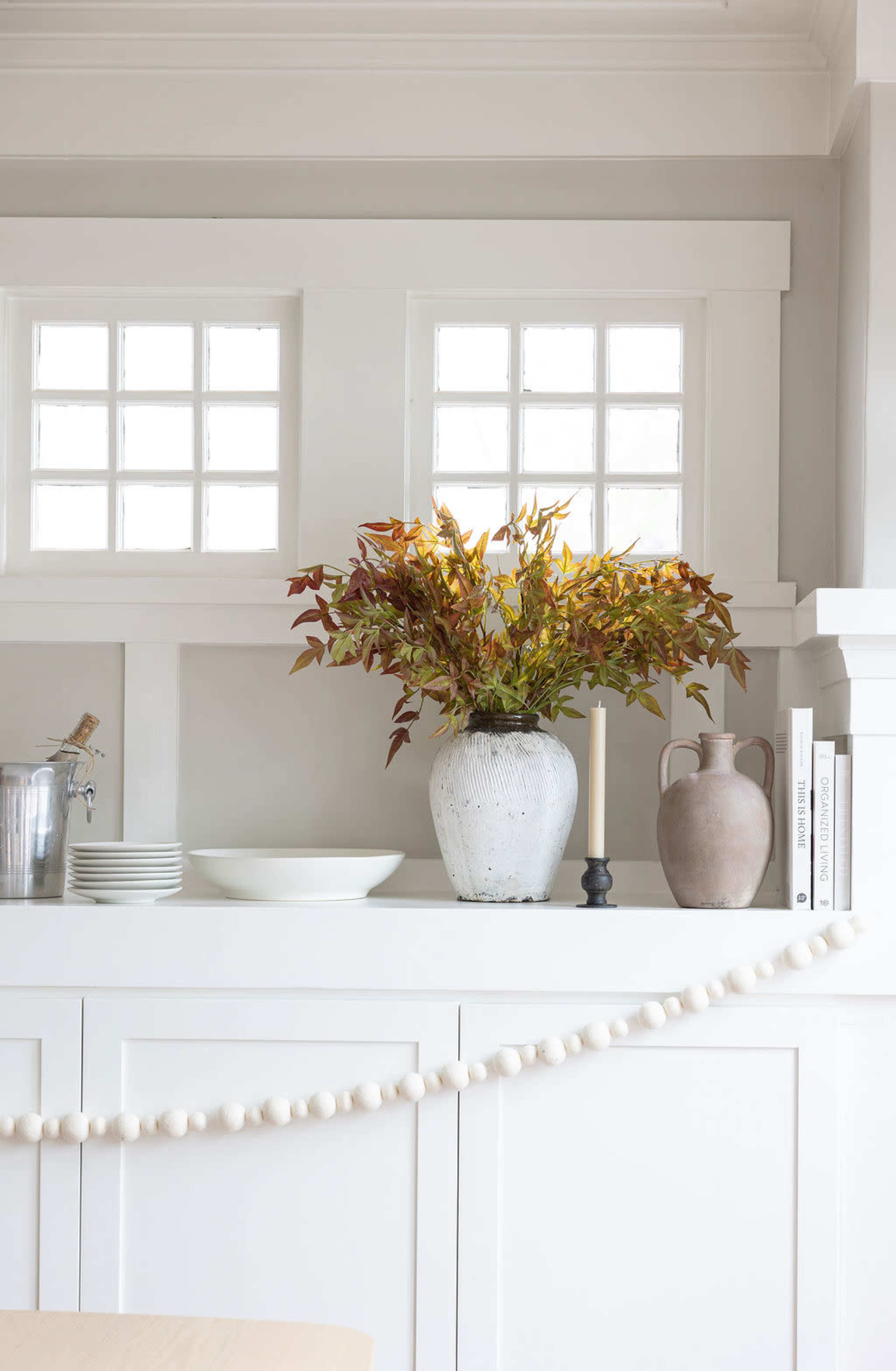 A decorated shelf features a vase of foliage, a candle holder, plates, a decorative pitcher, and a string of decorative beads.