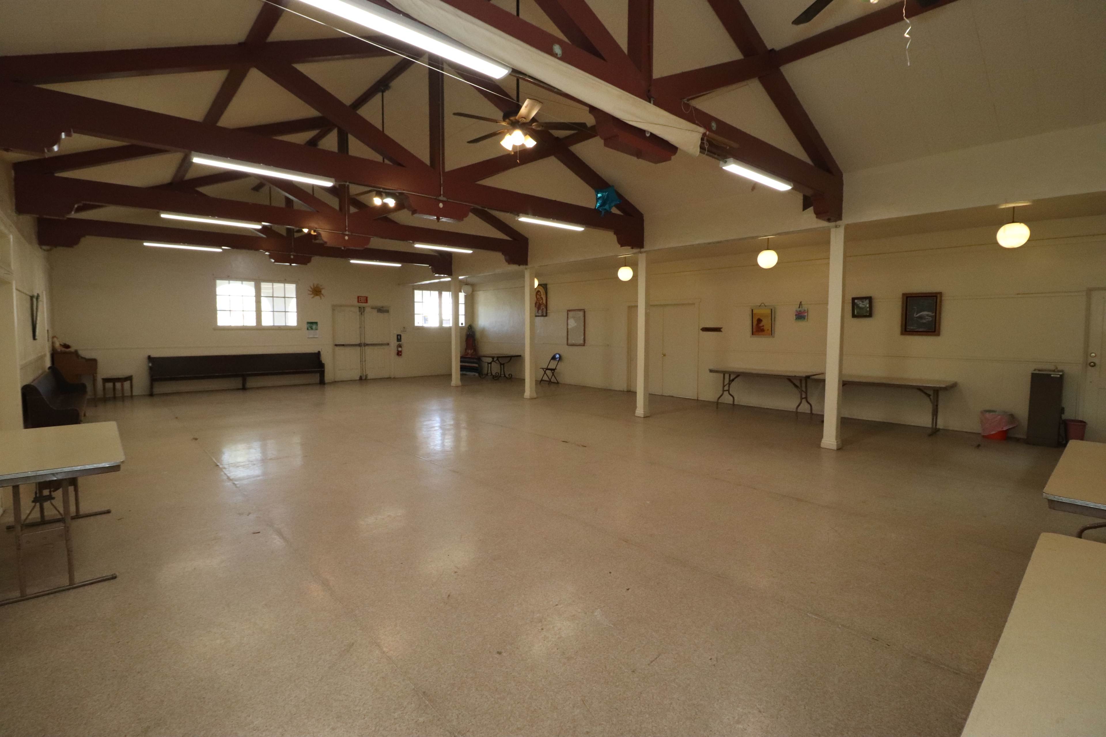 A spacious interior room with a high ceiling, featuring several tables and chairs along the walls, and large windows providing natural light.