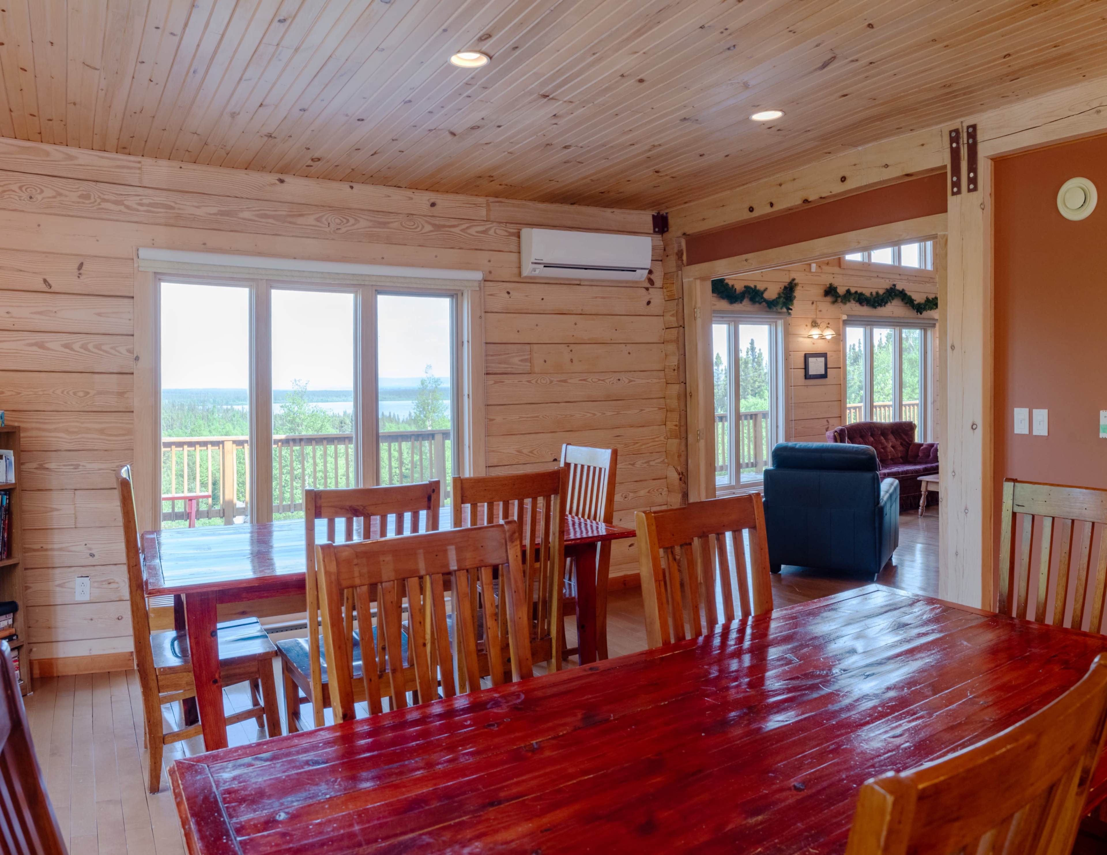 The image shows a rustic wooden interior with a dining area featuring a large table surrounded by chairs, and a view of a balcony overlooking a landscape through sliding glass doors.