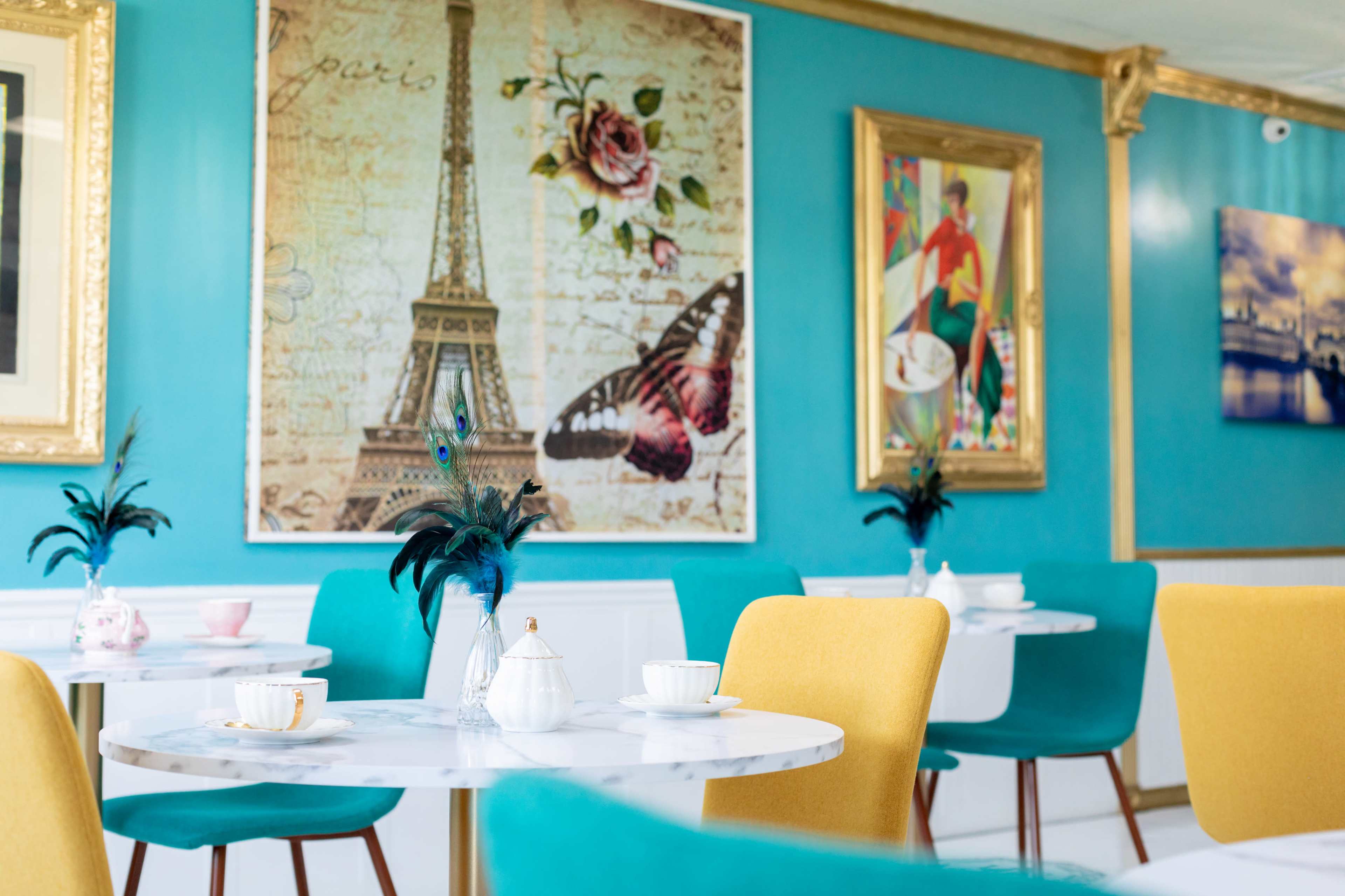 A brightly colored café with turquoise walls, marble tables set with tea cups, and framed artwork featuring an Eiffel Tower and butterflies.