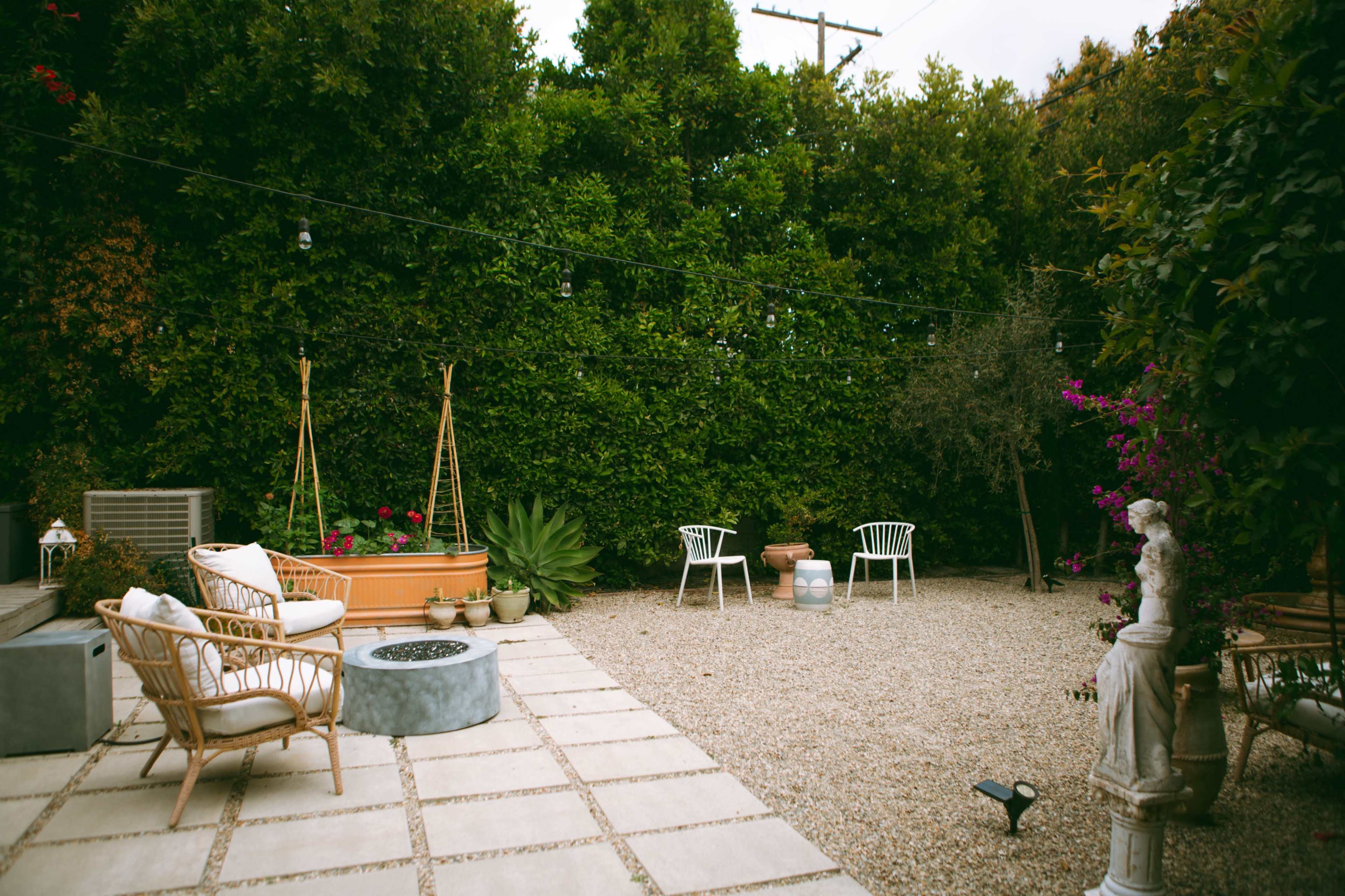 The image shows a landscaped garden area with a gravel surface, outdoor seating, and potted plants surrounded by lush greenery.