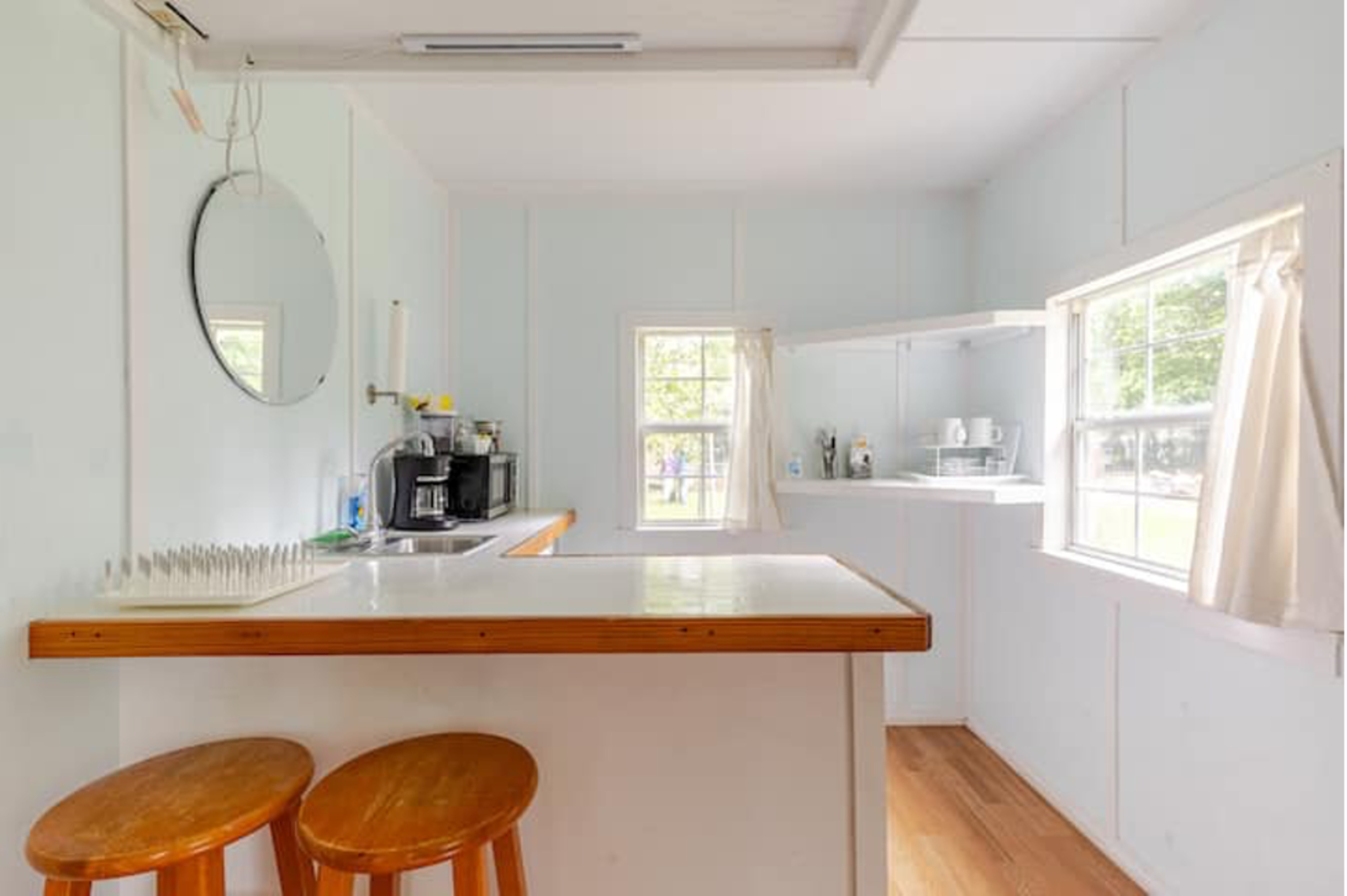 A light blue kitchen with a bar area featuring two wooden stools, a coffee maker, and a window with white curtains.