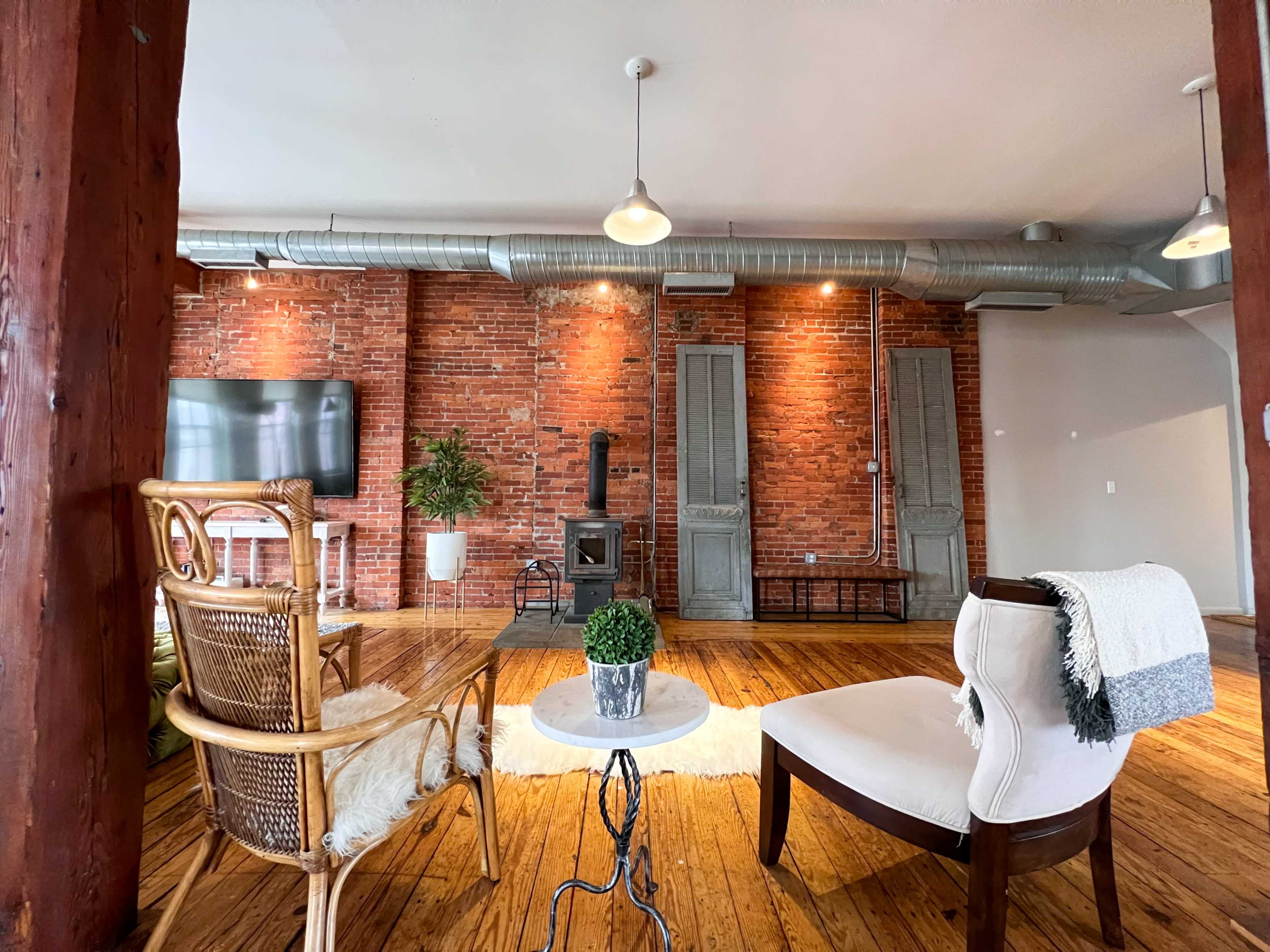 A modern living room with exposed brick walls, wooden flooring, and minimalistic furniture including a white chair, a wicker chair, and a small round table.