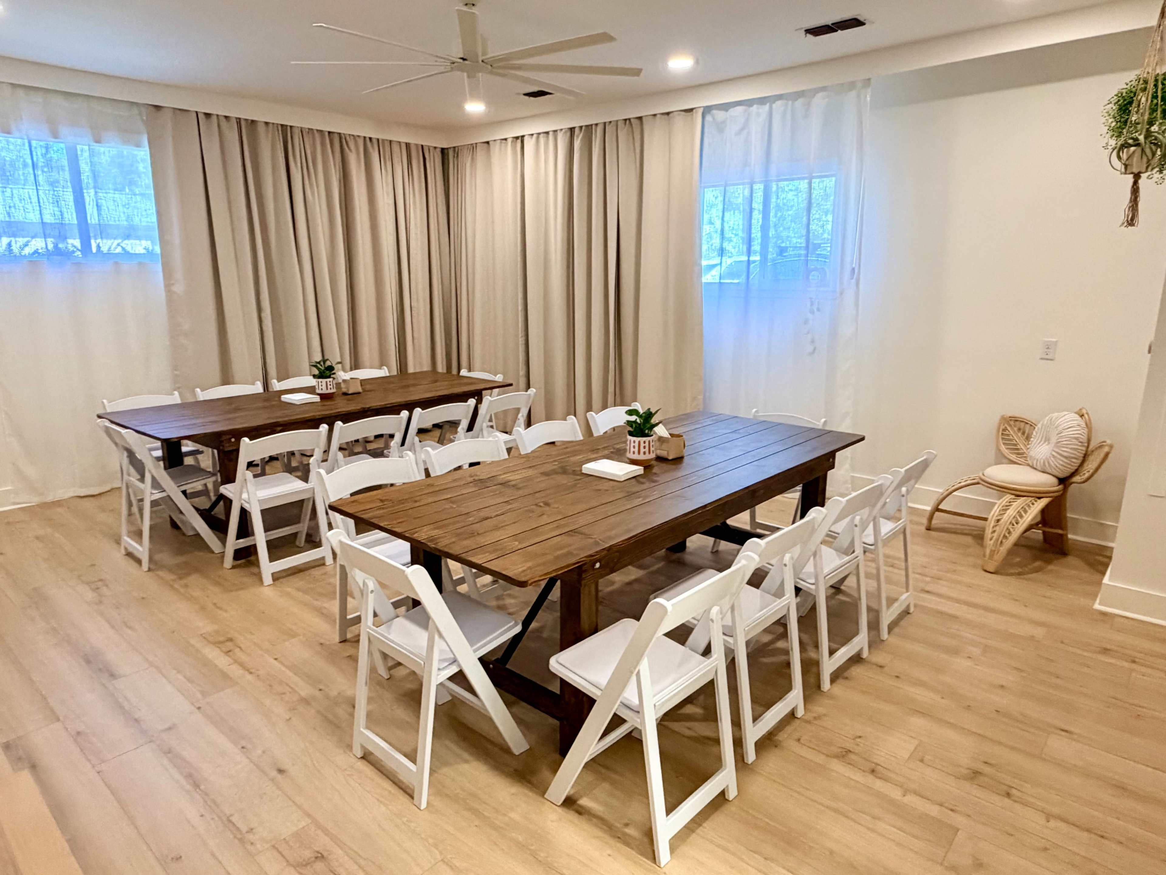 The image shows a well-lit dining area featuring two long wooden tables surrounded by white folding chairs and soft cream curtains on the windows.