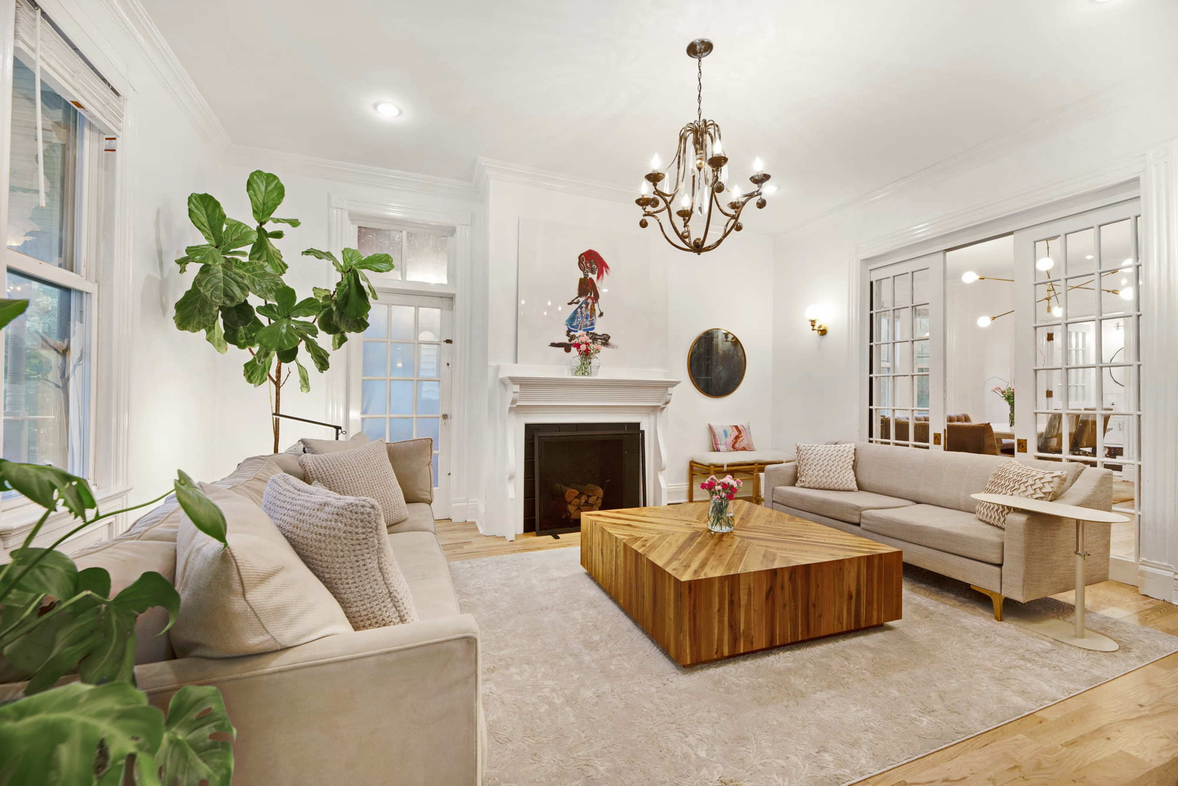 A bright living room featuring two sofas, a large wooden coffee table, a fireplace, and large windows with a view of indoor plants.