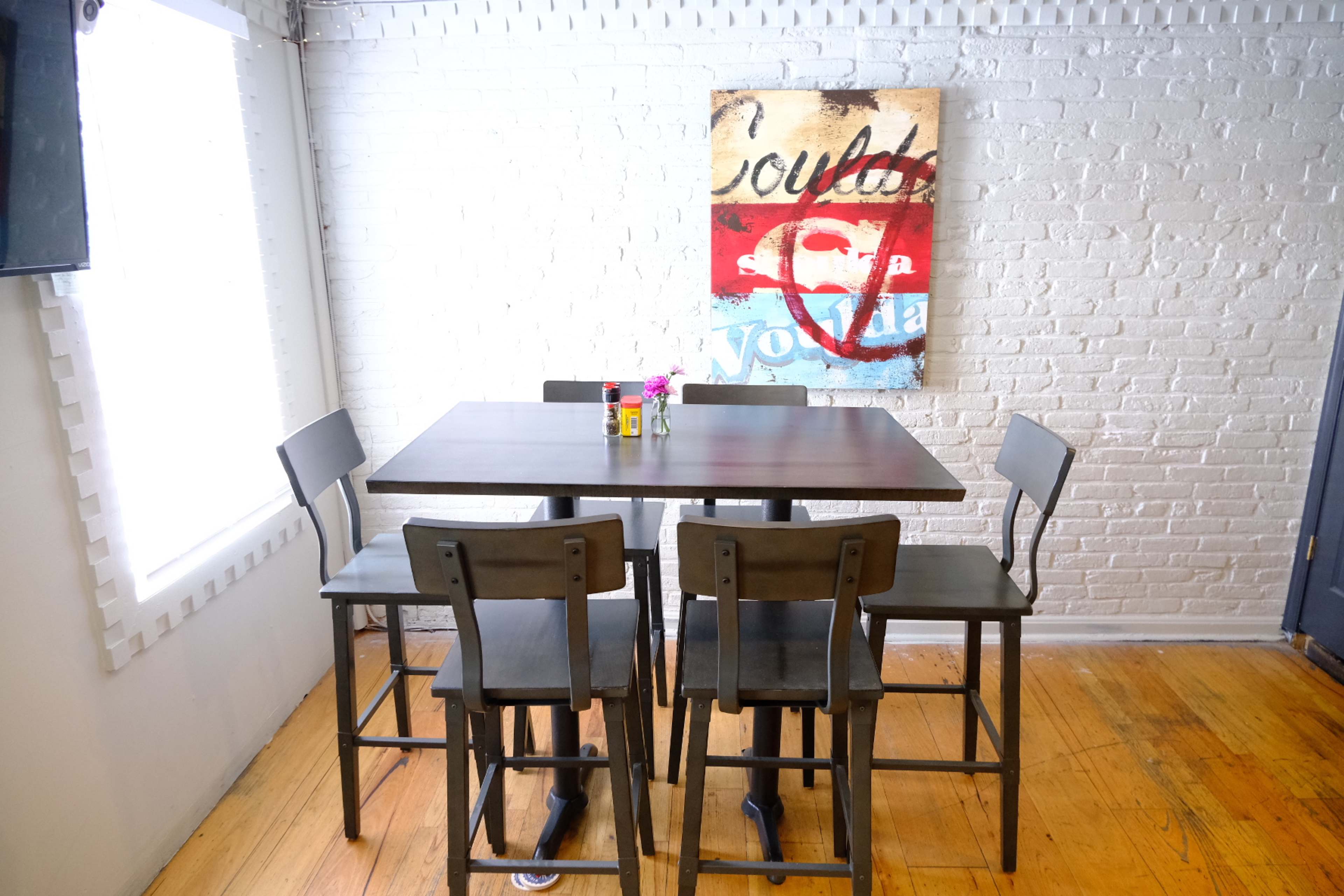 A wooden table surrounded by four black high-backed chairs sits in a room with white brick walls and a colorful piece of art hanging above.