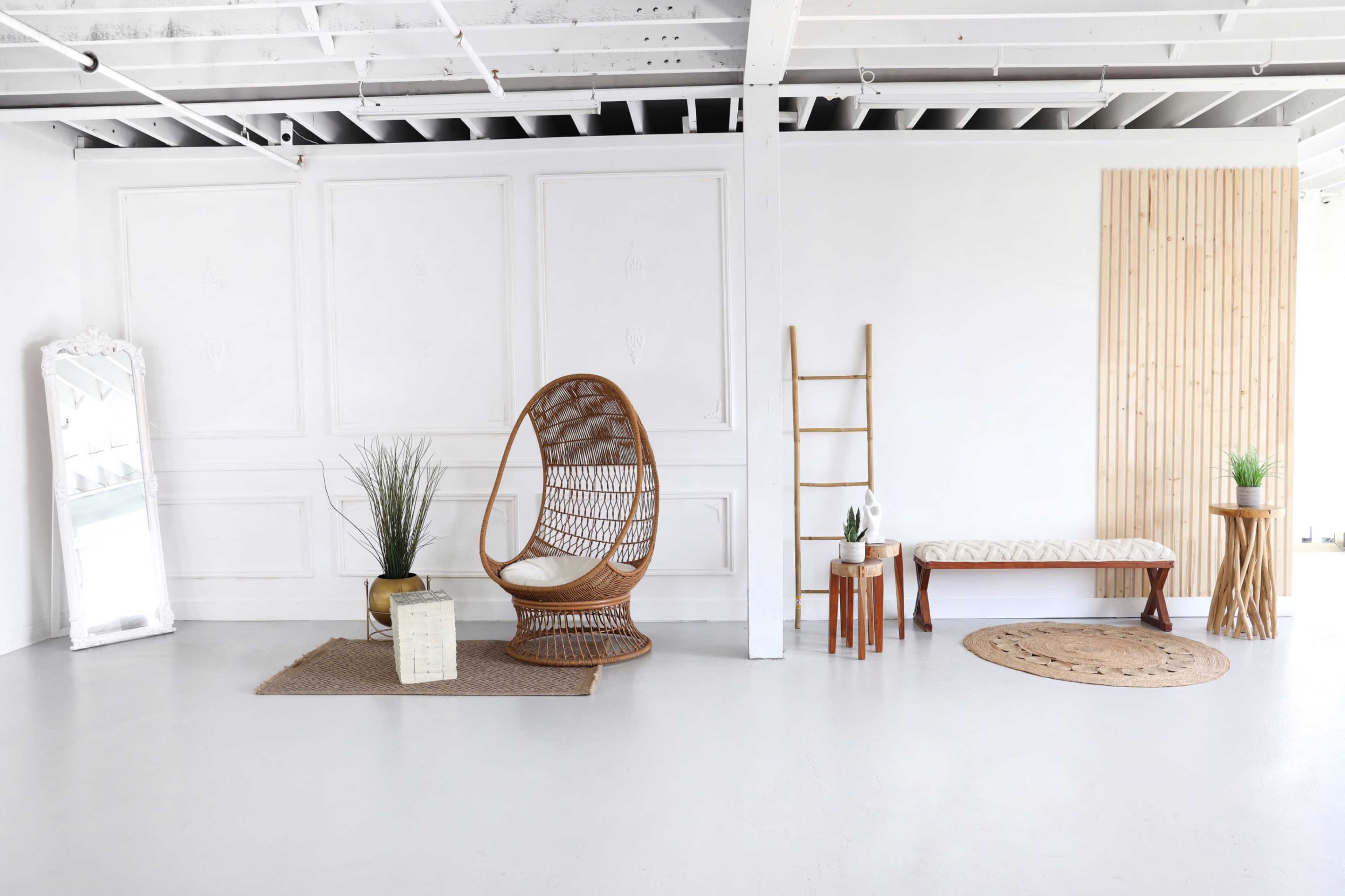 A minimalist interior space featuring a rattan chair, a mirror, various decorative plants, and several wooden furniture pieces against a white wall.