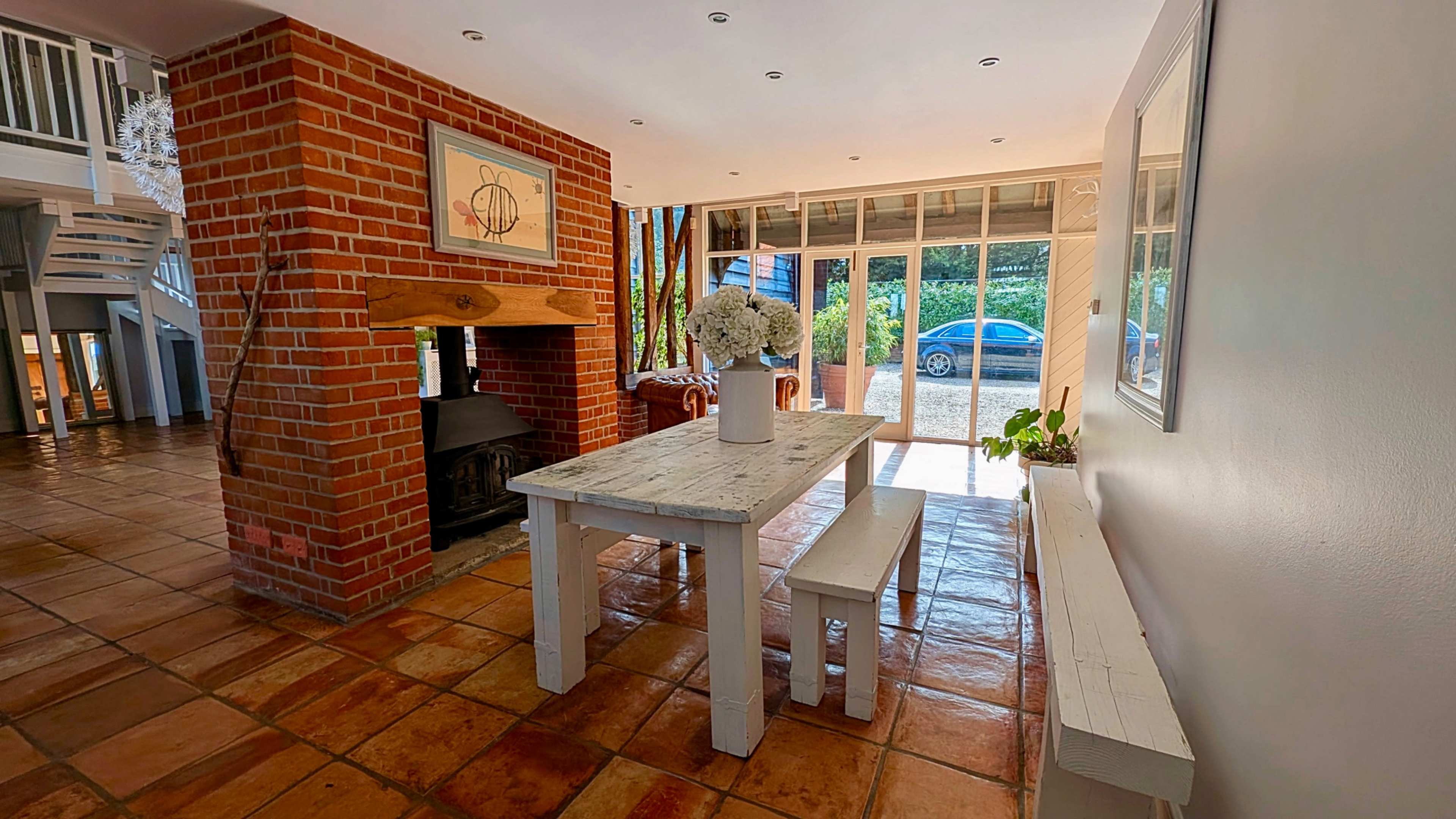 The image shows a spacious dining area with a wooden table and benches, a brick fireplace, and large windows offering natural light.