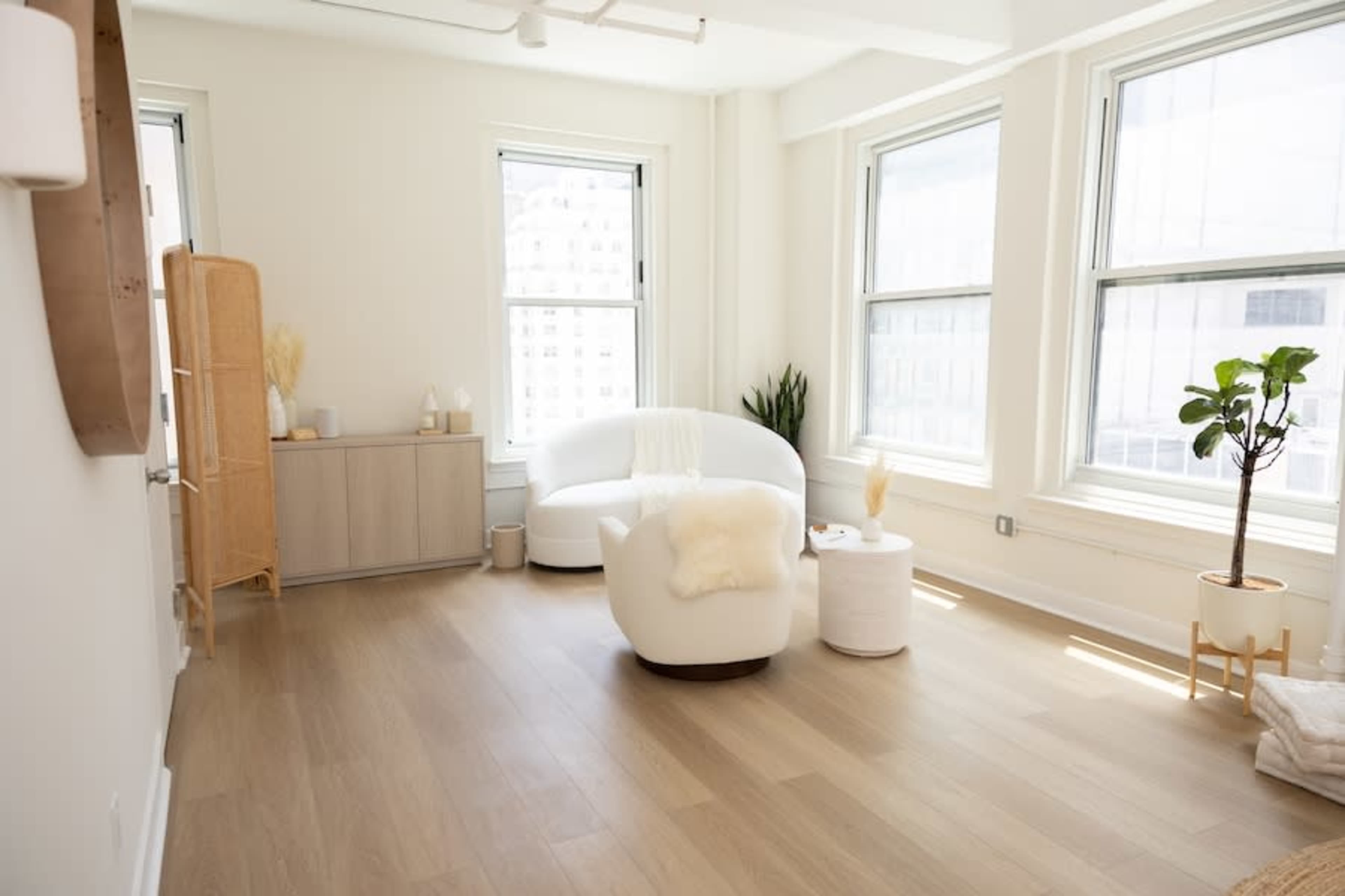 The scene shows a bright, minimalist living space featuring a white sectional sofa, a wooden cabinet, and large windows letting in natural light.