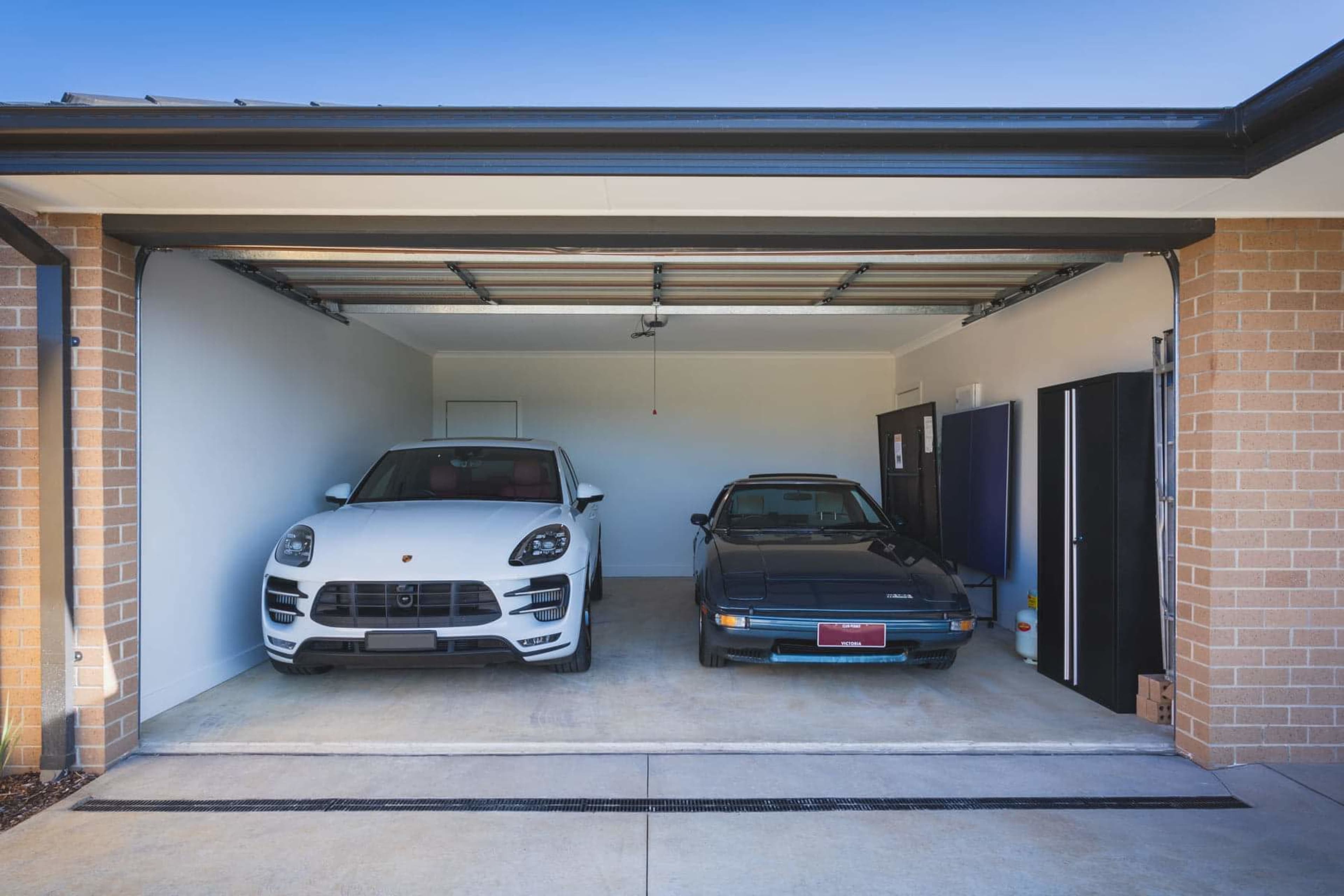 A white Porsche Macan and a black sports car are parked inside a two-car garage with a concrete floor and beige brick walls.