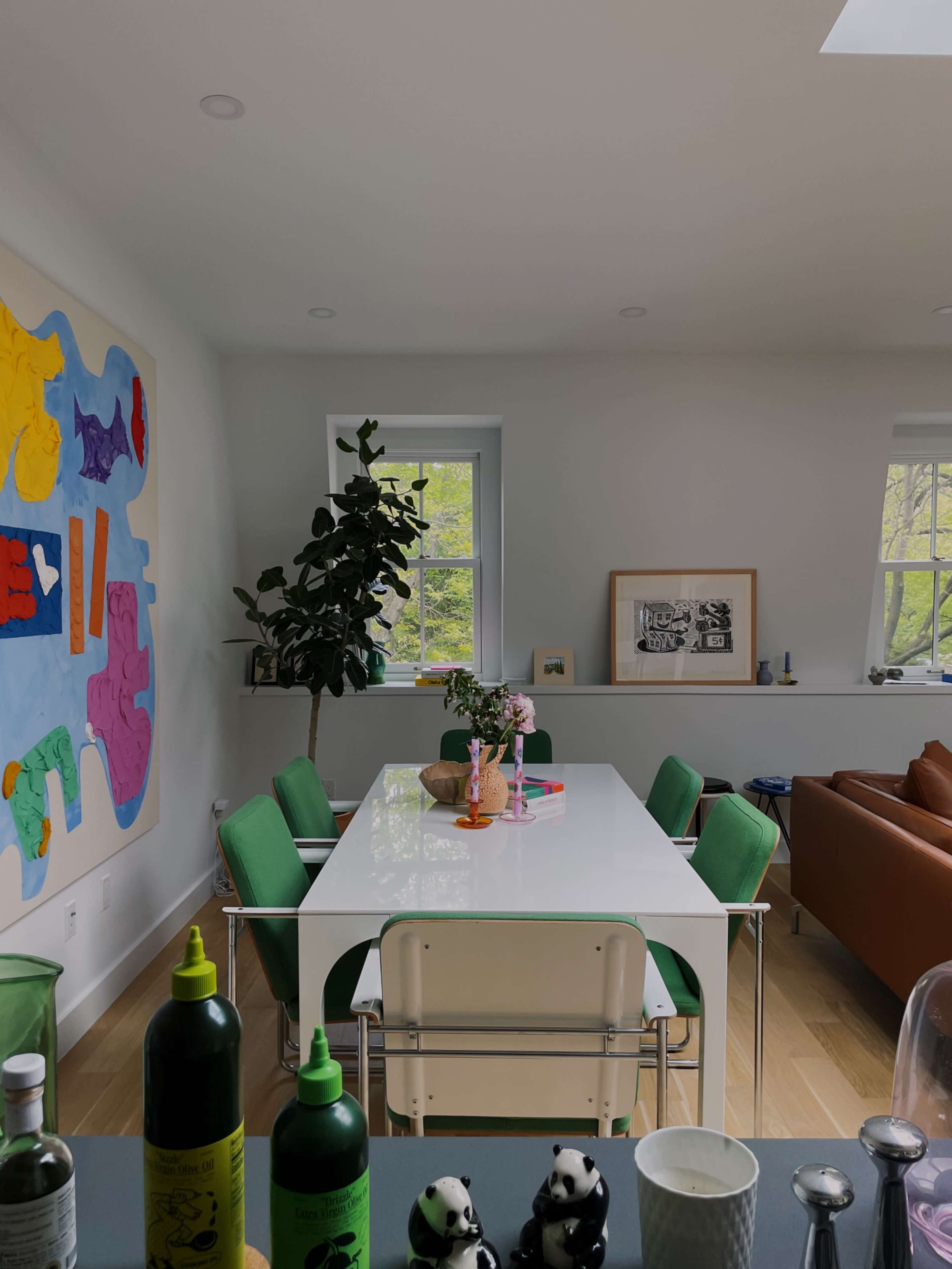 The image shows a modern dining area featuring a white table, green chairs, a large colorful wall decoration, and a window view with plants.