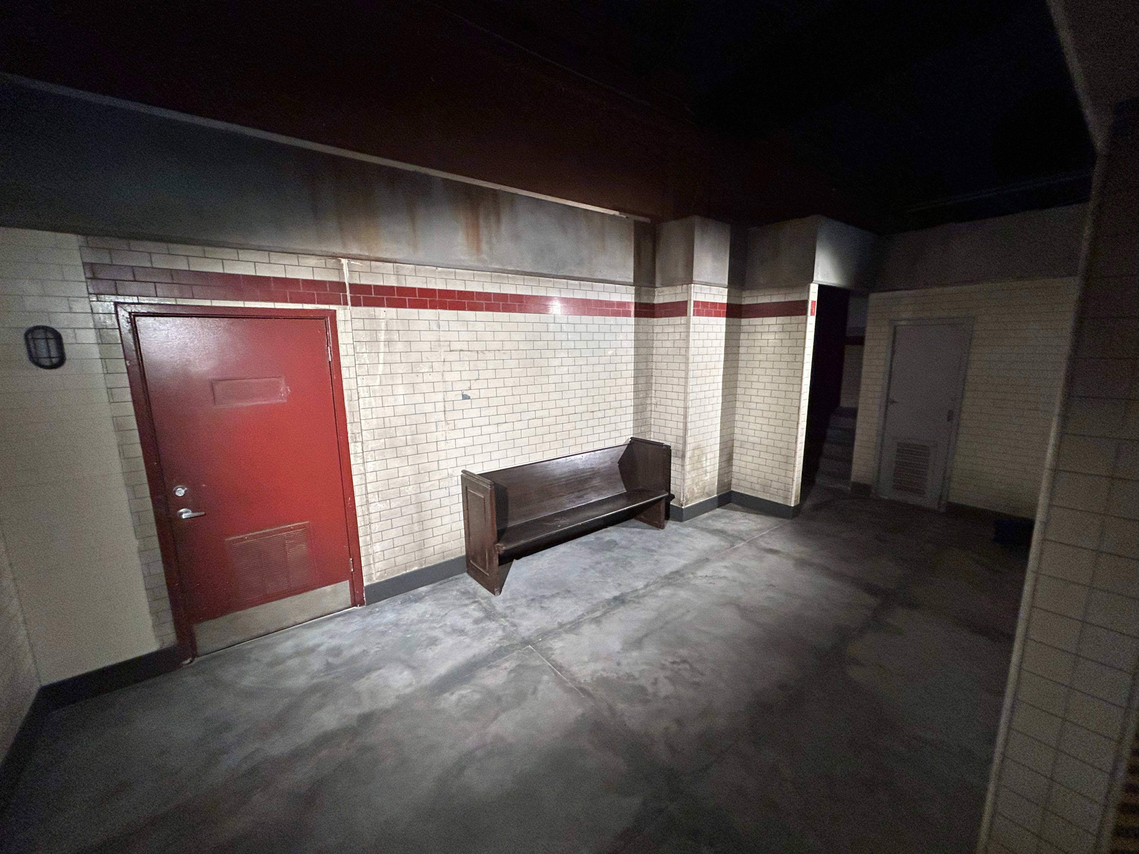 The image shows a dimly lit hallway with a wooden bench and red-accented walls, featuring brick tiles and a door at one end.