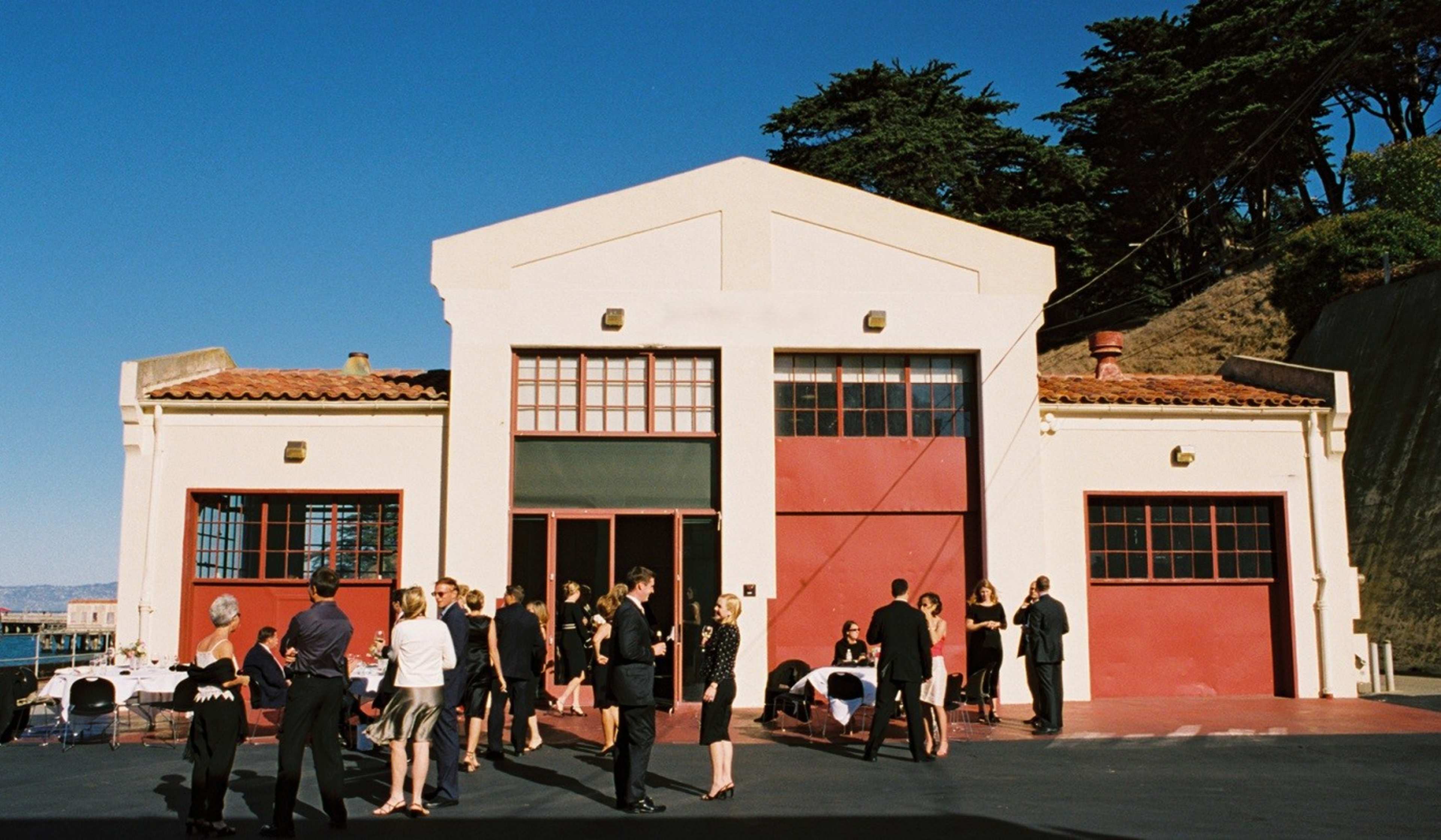 A group of people in formal attire socializes in front of a large building with red doors, near a waterfront.
