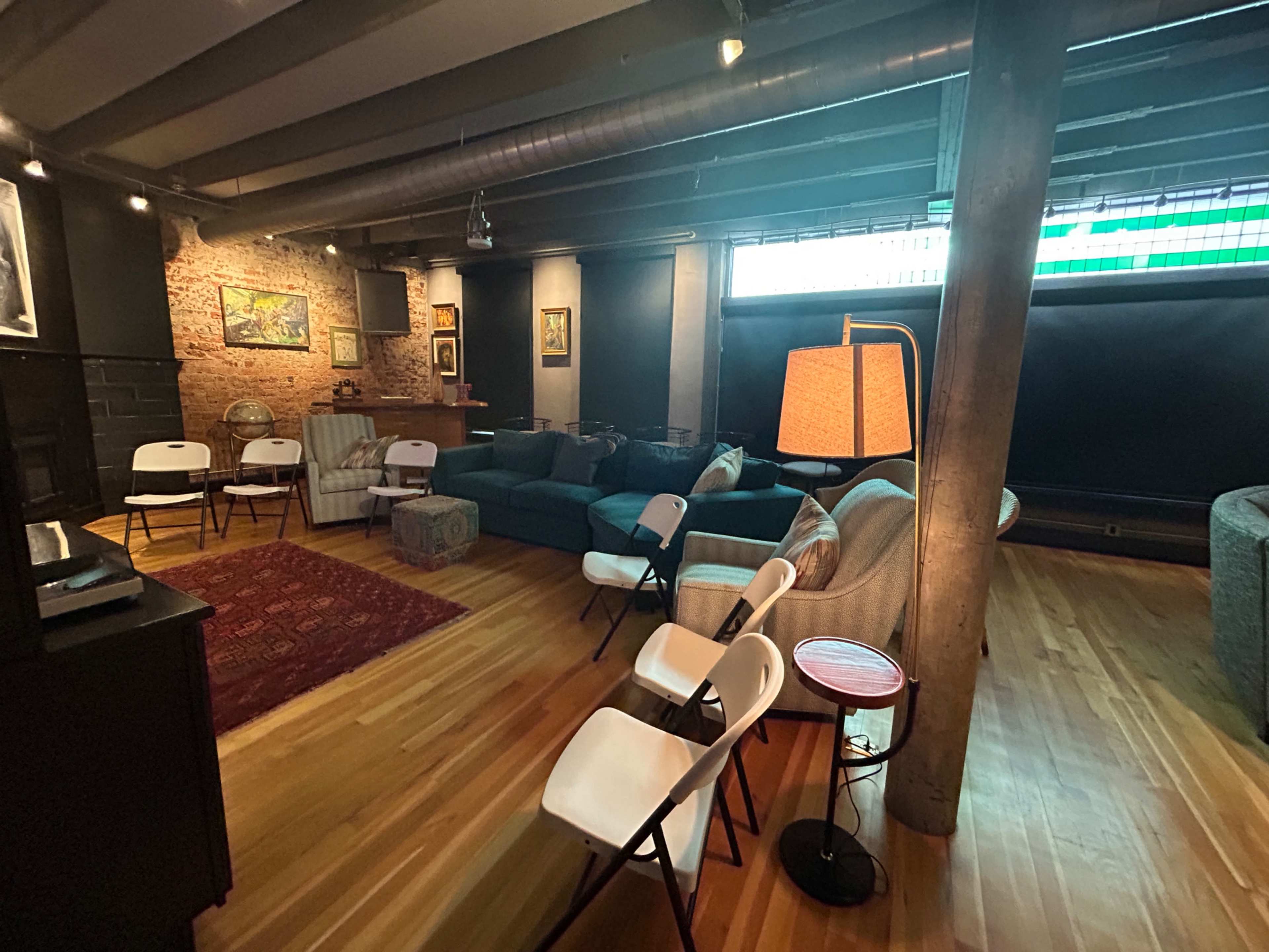 The image shows a spacious living area with a combination of seating options including sofas and folding chairs, wooden flooring, and exposed brick walls, illuminated by various light sources.