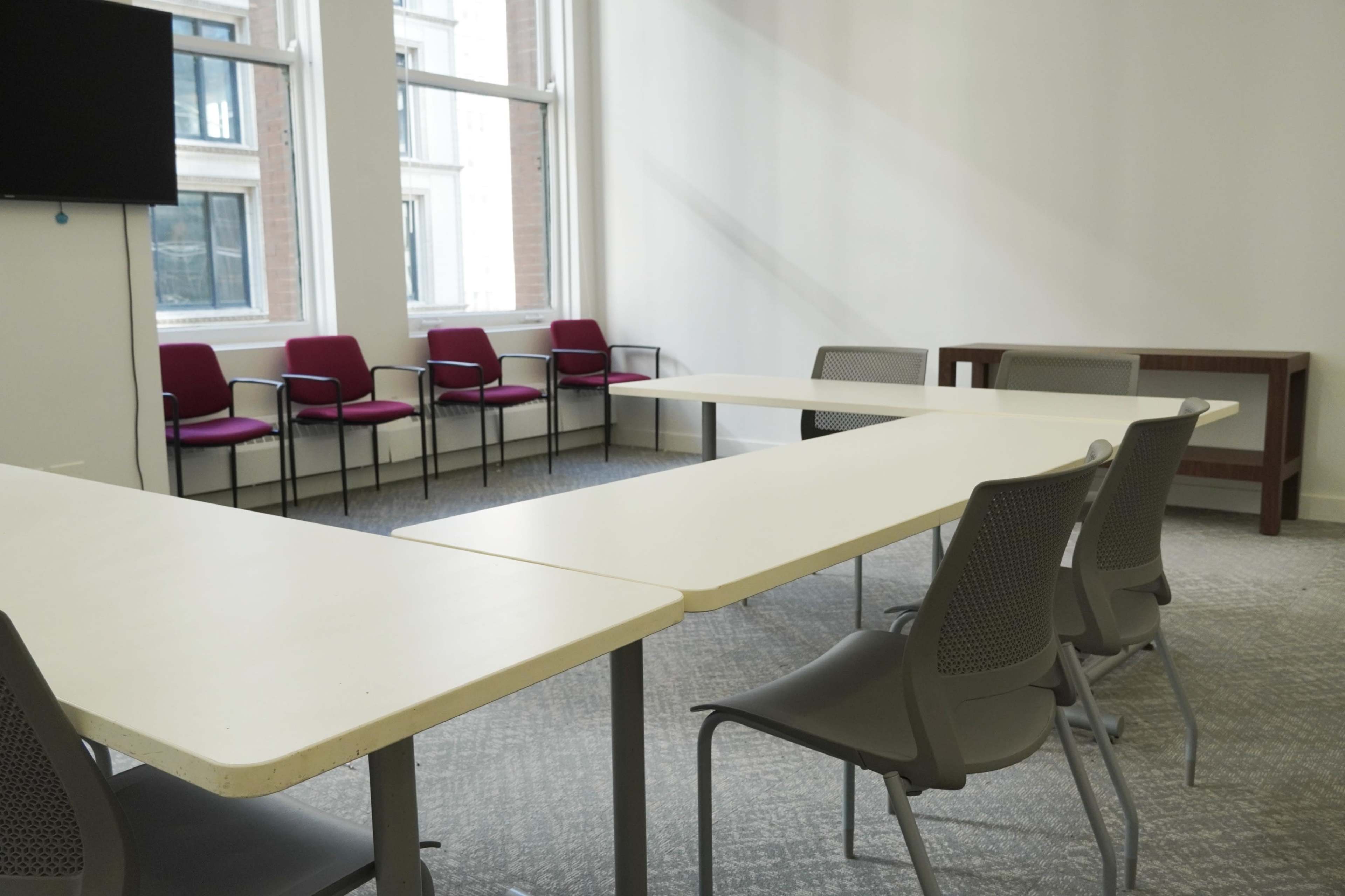 A conference room features a large table surrounded by gray chairs, with additional red chairs against the wall and windows letting in natural light.