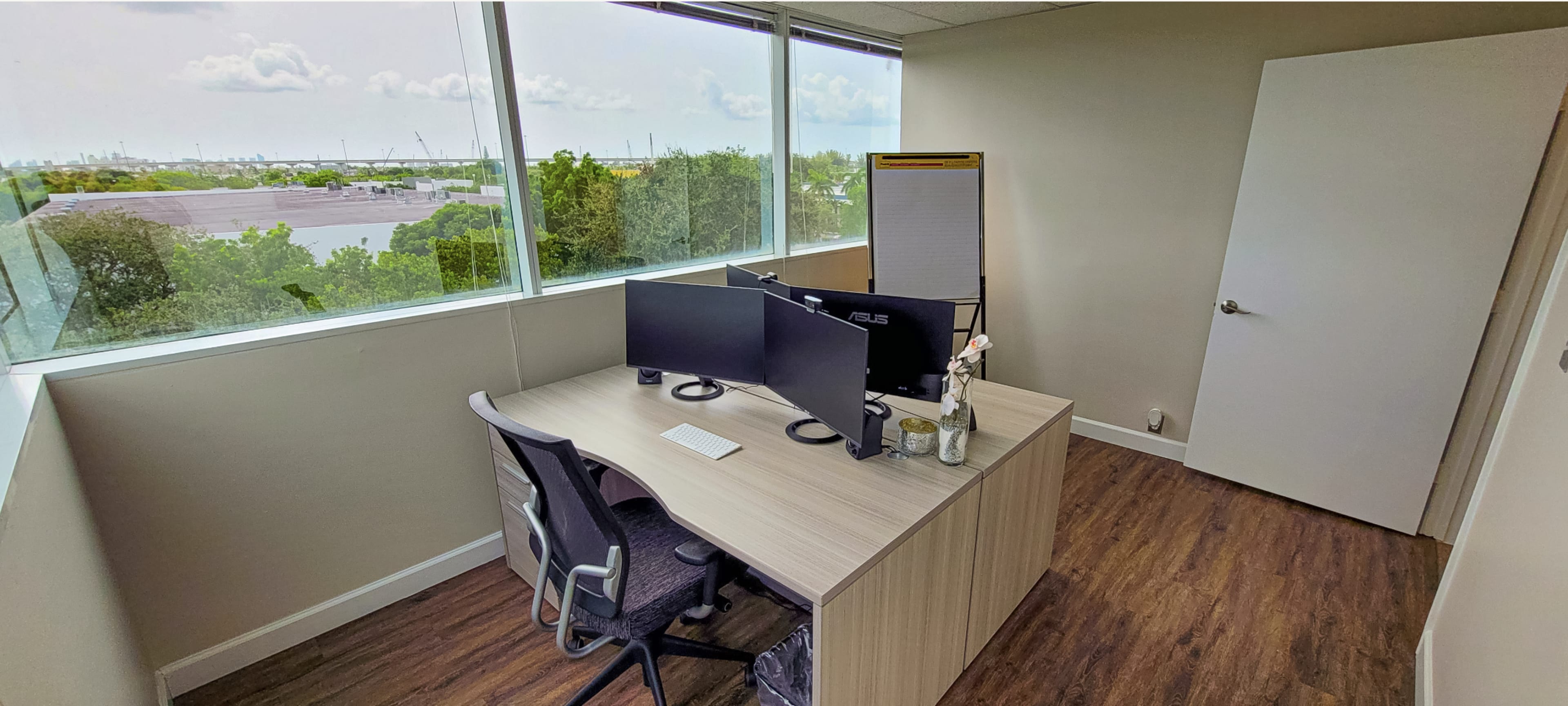 A minimalist office space with two computer monitors on a light wooden desk, a chair, and large windows providing a view of greenery and wind turbines outside.