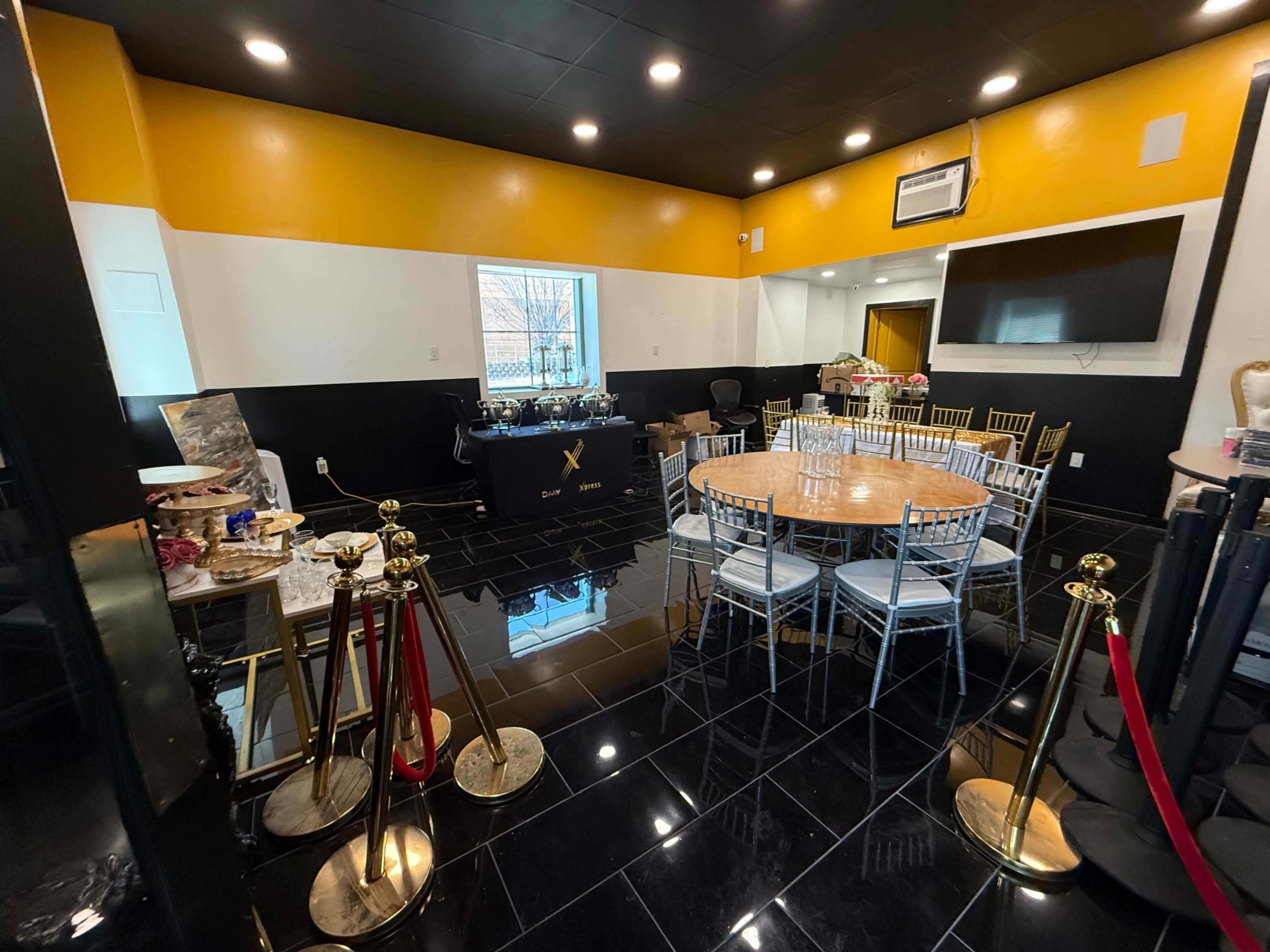 The image shows a brightly colored event space featuring a round wooden table surrounded by white chairs, with a service area in the background displaying snacks and beverages.