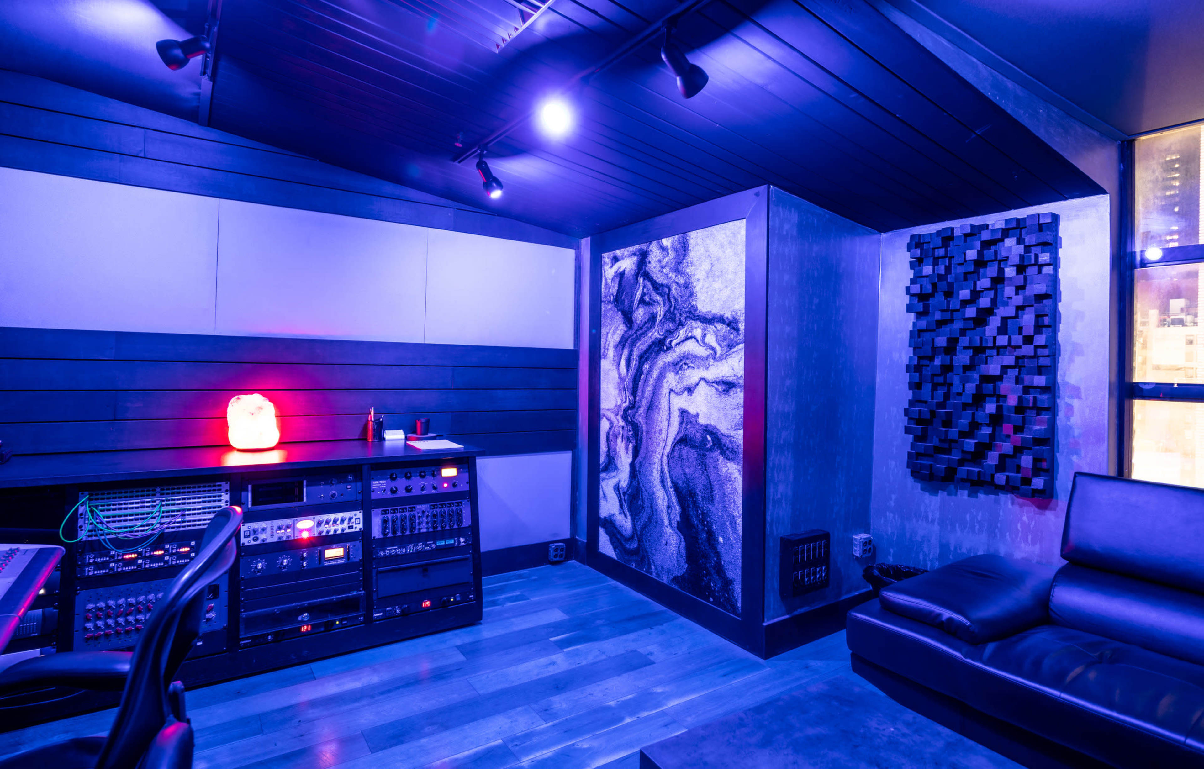 A sound studio features dark walls, a sleek sofa, and various audio equipment, illuminated by purple lighting.