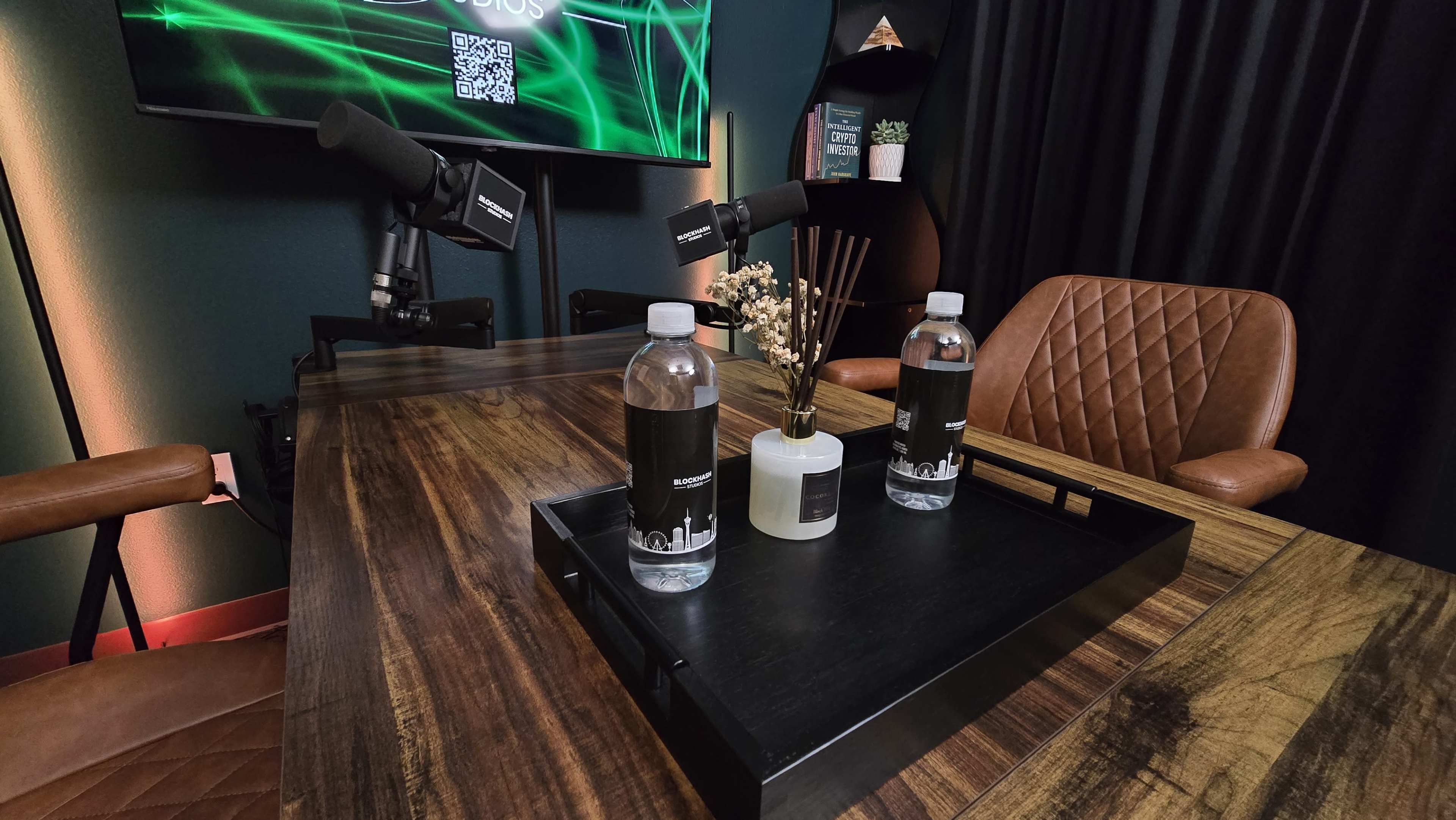 A wooden conference table with two water bottles and a small decorative vase is set in a recording studio featuring microphones and a digital screen in the background.