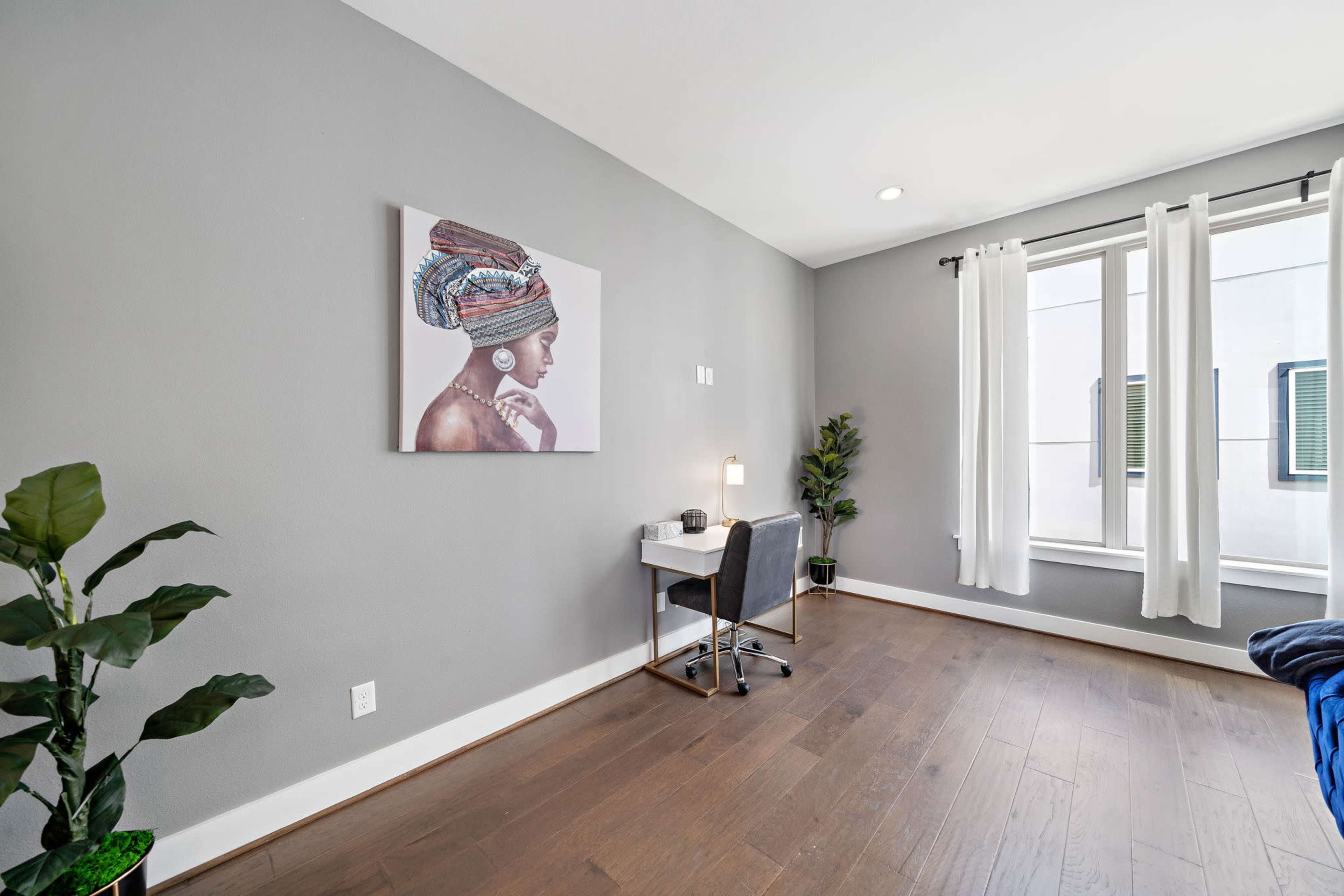 A modern workspace with a small desk and chair next to a window, featuring a wall art of a woman with a colorful head wrap and a potted plant in the corner.