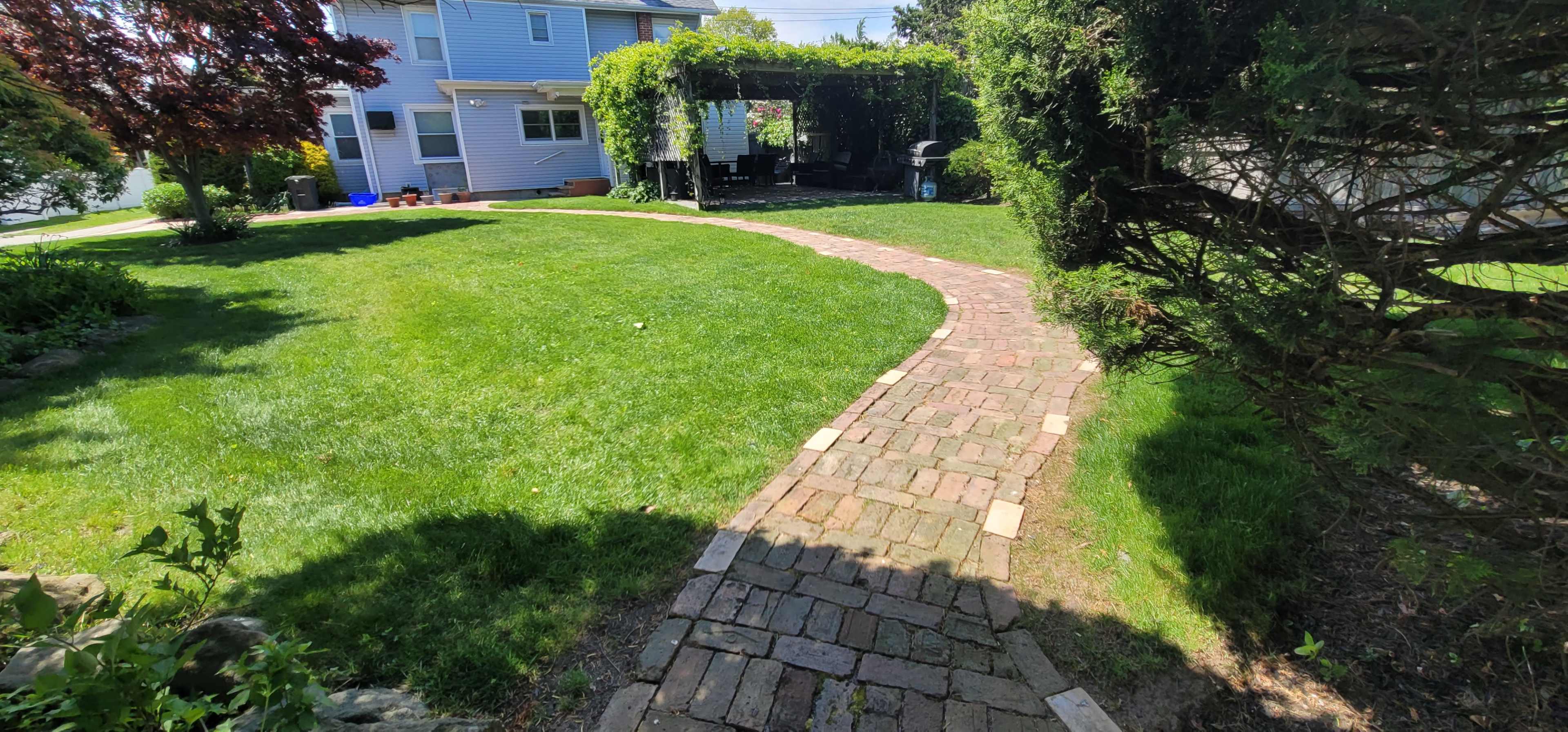 A curved brick pathway leads through a green lawn, bordered by hedges and a shaded patio area beside a two-story house.