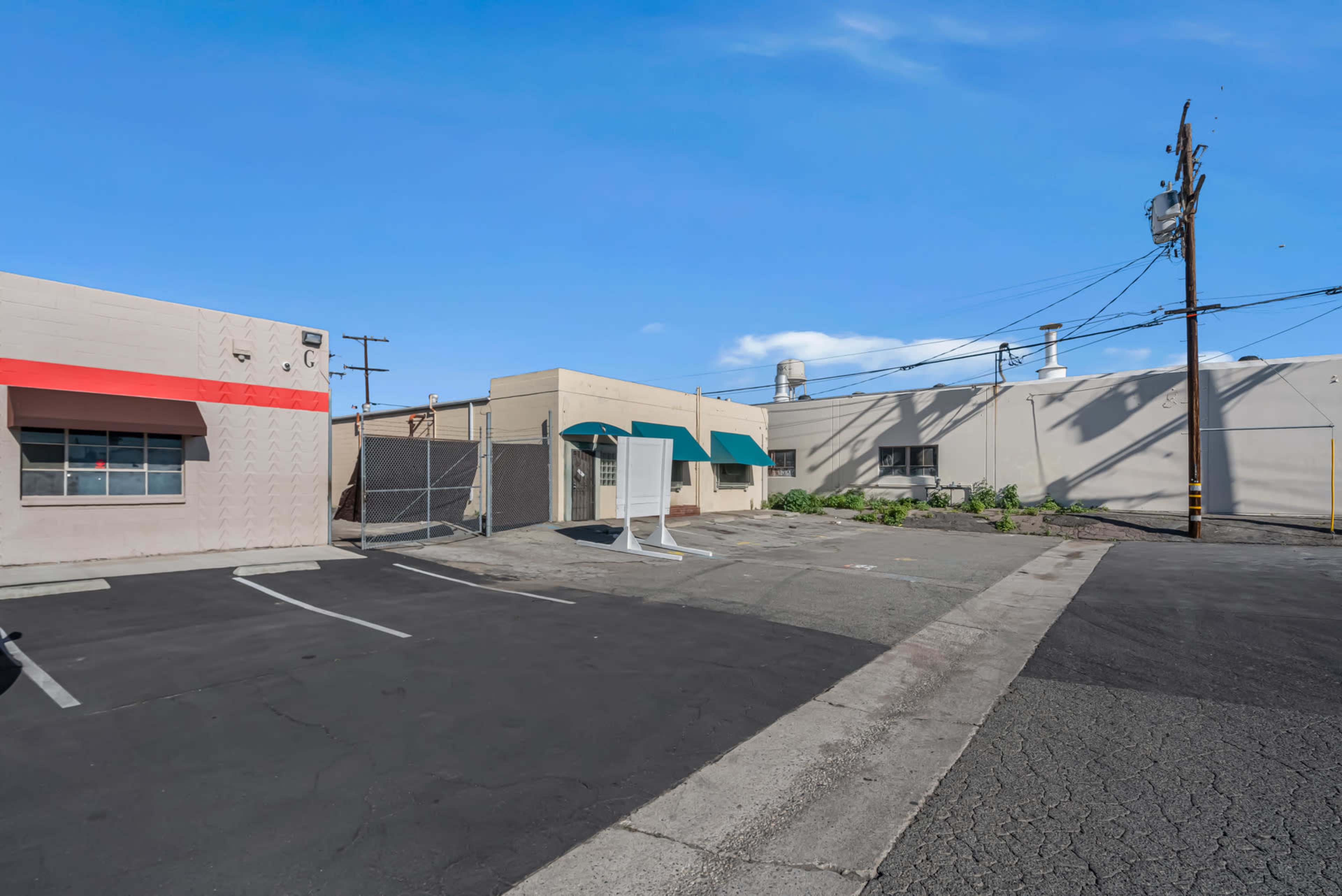 The image shows a vacant parking lot adjacent to several commercial buildings with visible overhead power lines and a clear blue sky.