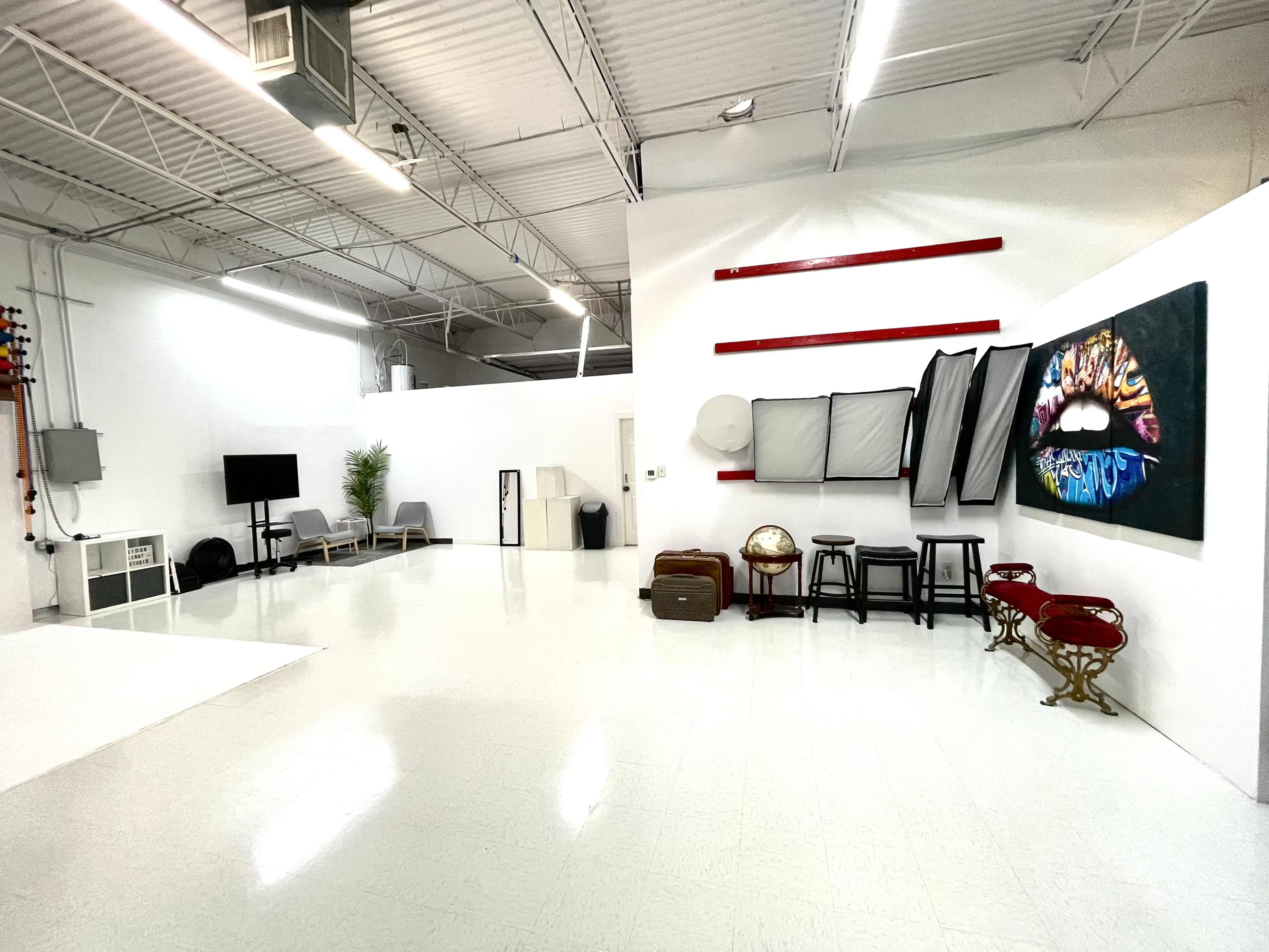 A spacious, well-lit studio with white walls, a polished floor, and various photography equipment, including softboxes and a large canvas featuring colorful lips.