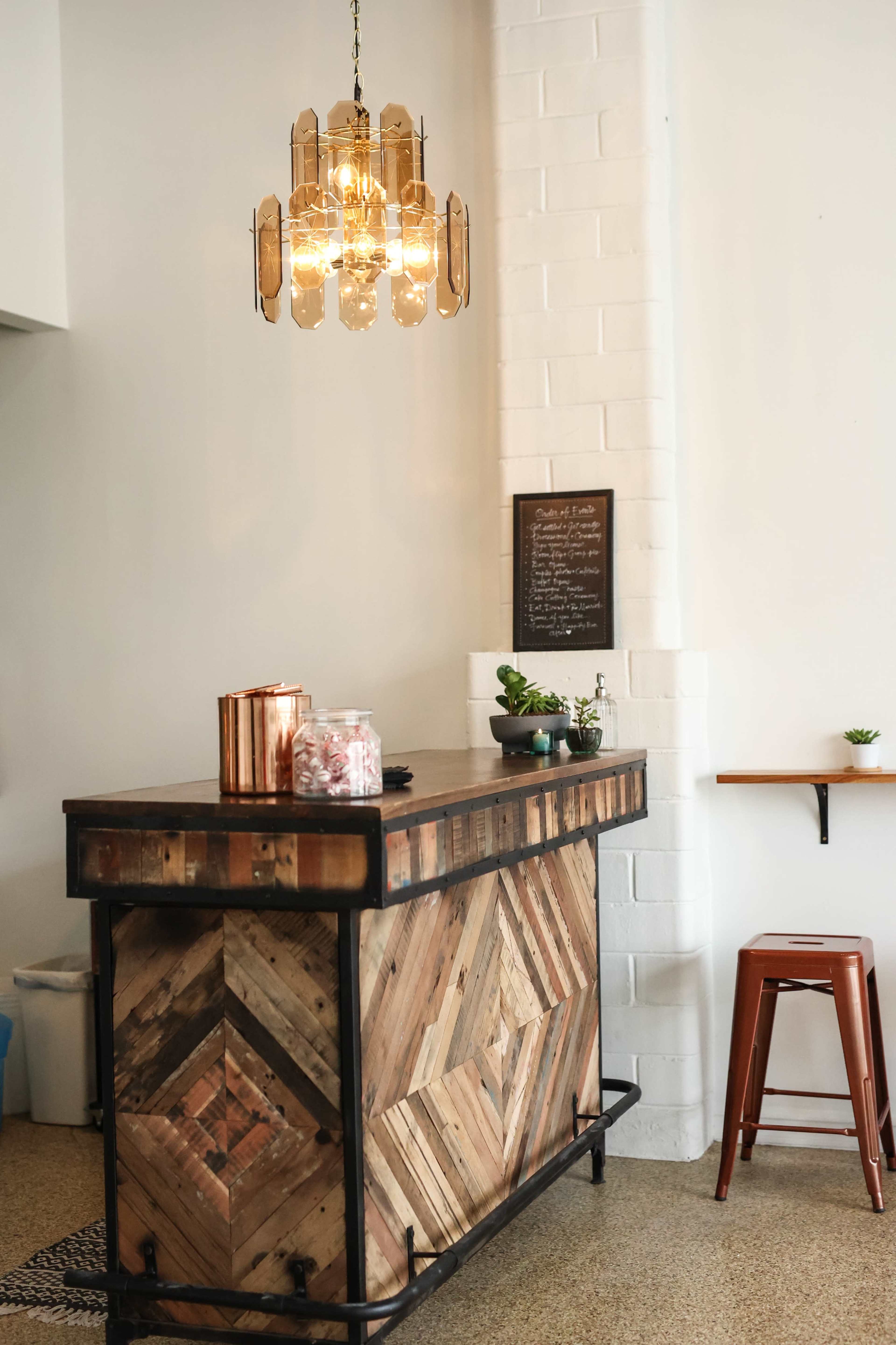 The image shows a wooden bar with a geometric design, a hanging light fixture, and a small menu board on the wall.