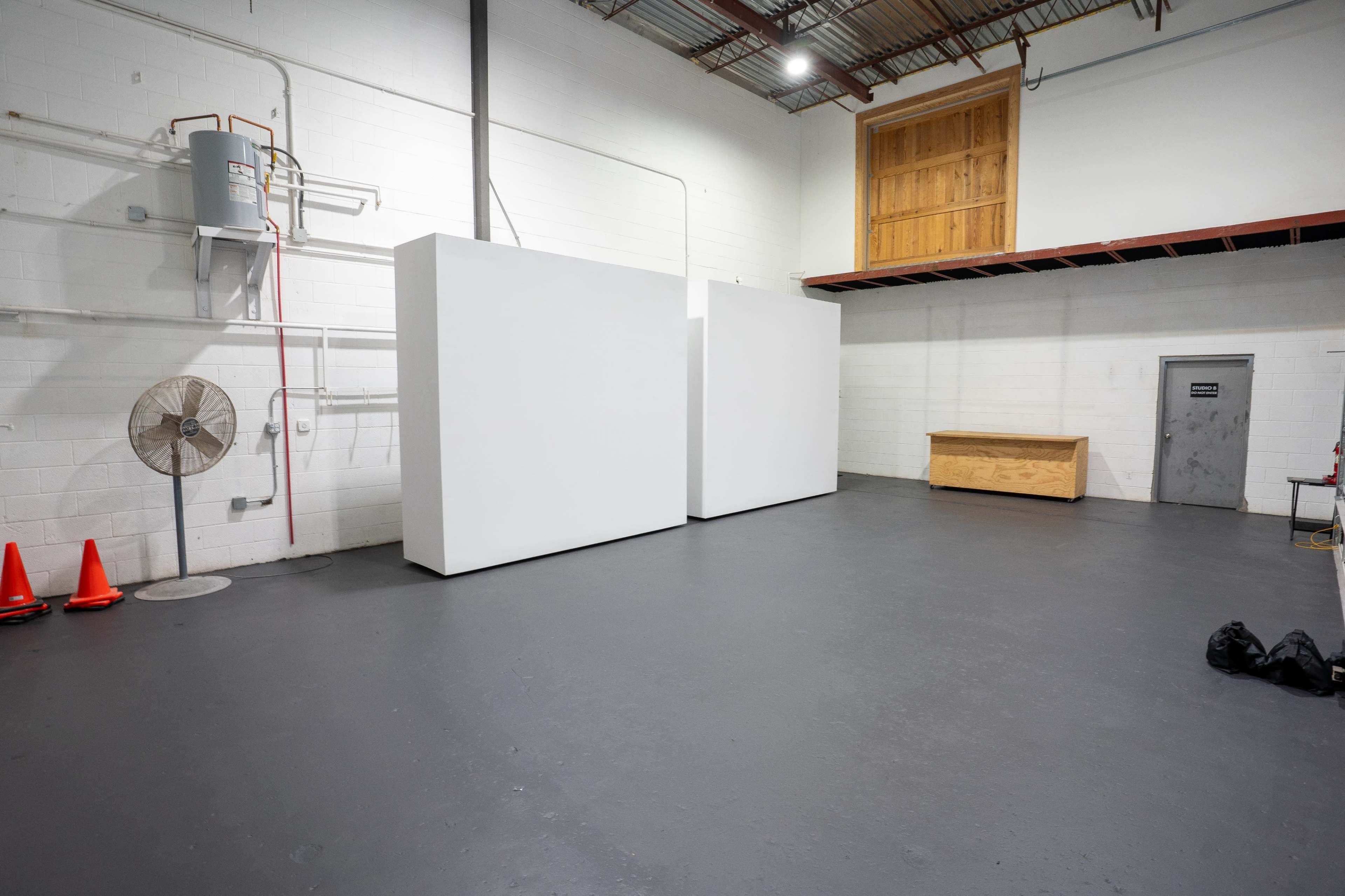 The image depicts a spacious, empty industrial room with two large white structures, a wooden storage chest, a fan, and orange cones on the floor.
