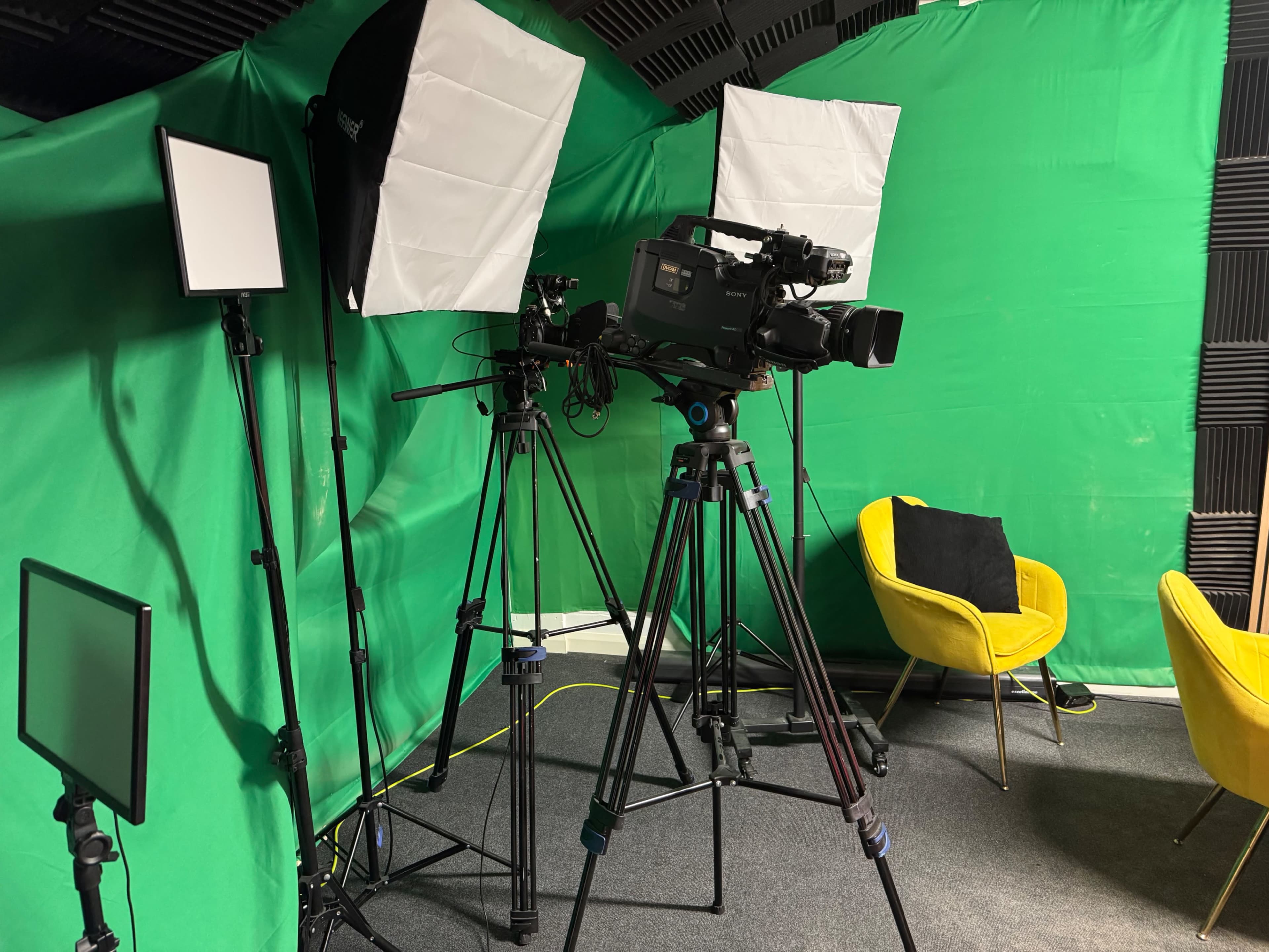 The image shows a studio setup with green backdrop, cameras on tripods, lighting equipment, and yellow chairs.