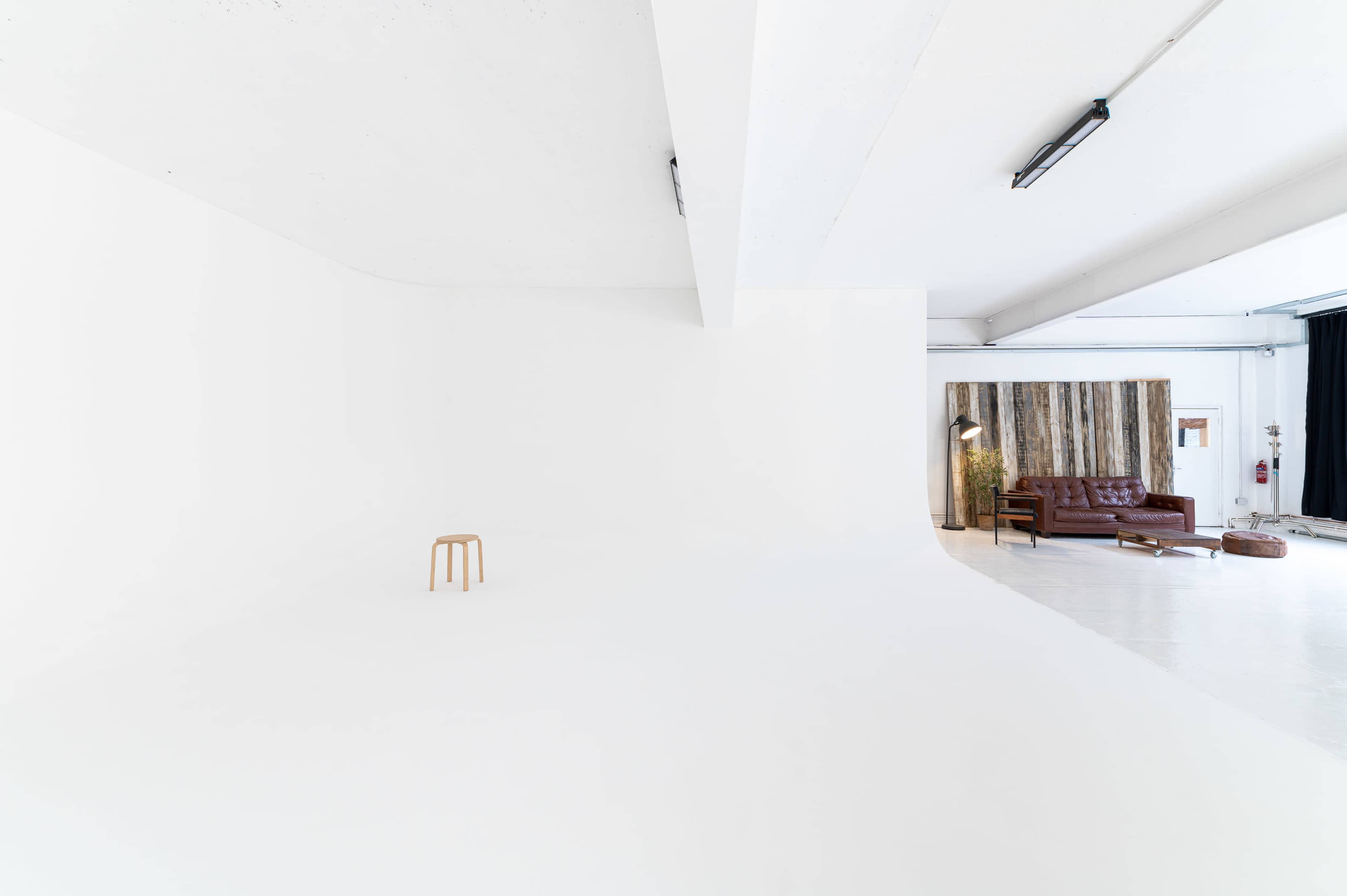 A minimalist studio space with a single wooden stool, a brown leather sofa, and a backdrop of vertical wooden planks.