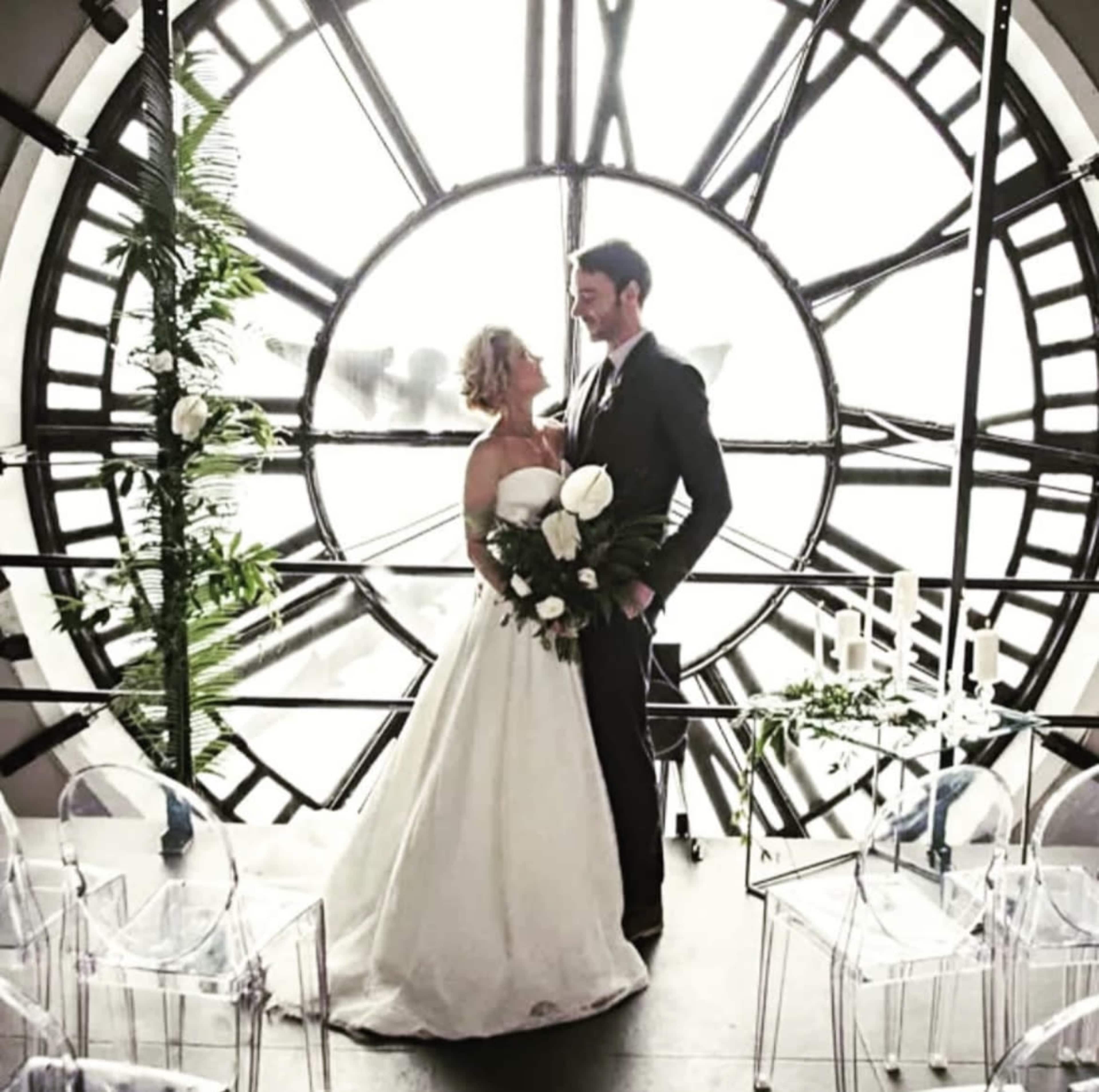 A couple stands together under a large clock face, framed by greenery and transparent chairs, as they prepare to exchange vows.